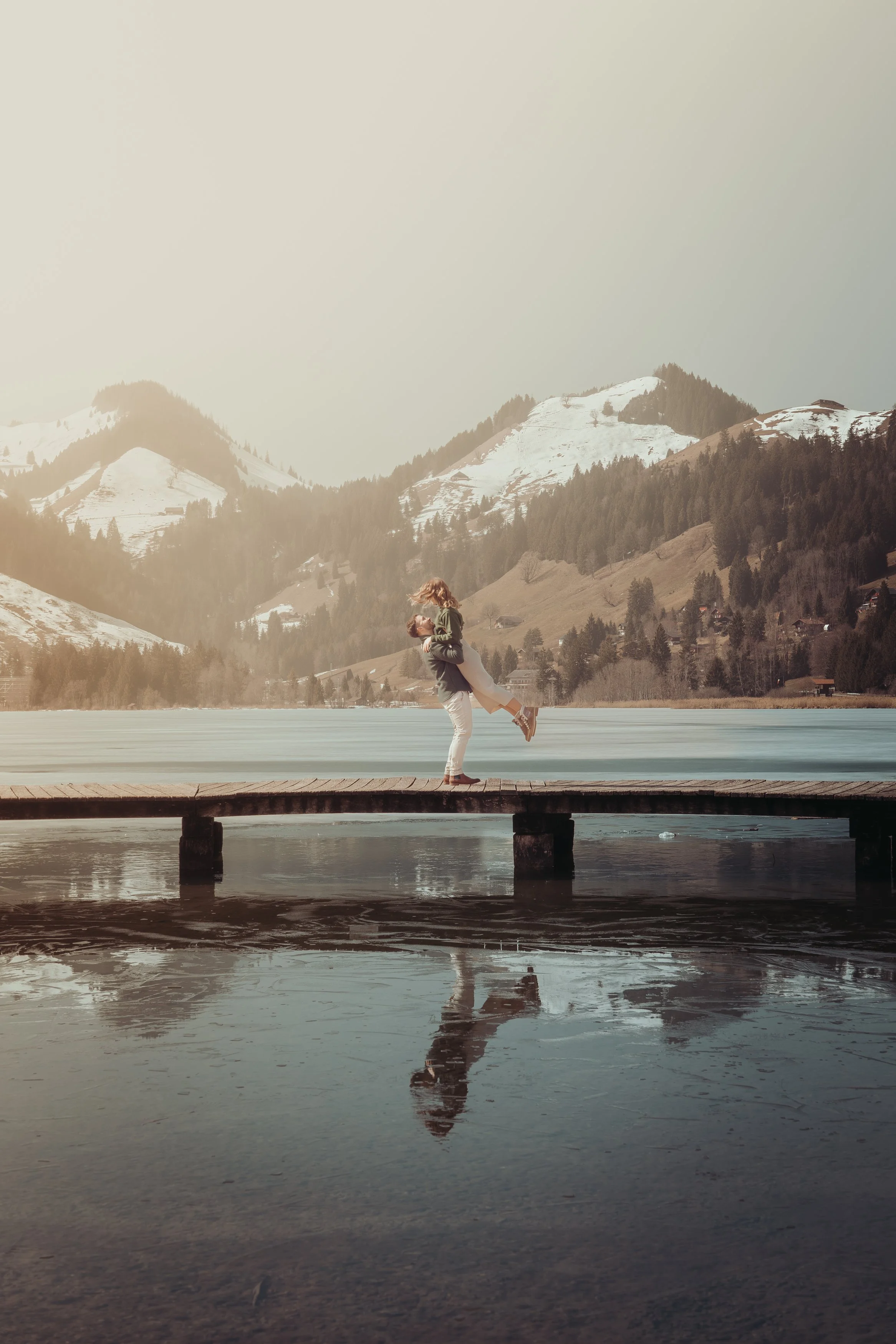 Un couple danse sur un pont en bois au bord d'un lac, avec des montagnes enneigées en arrière-plan.