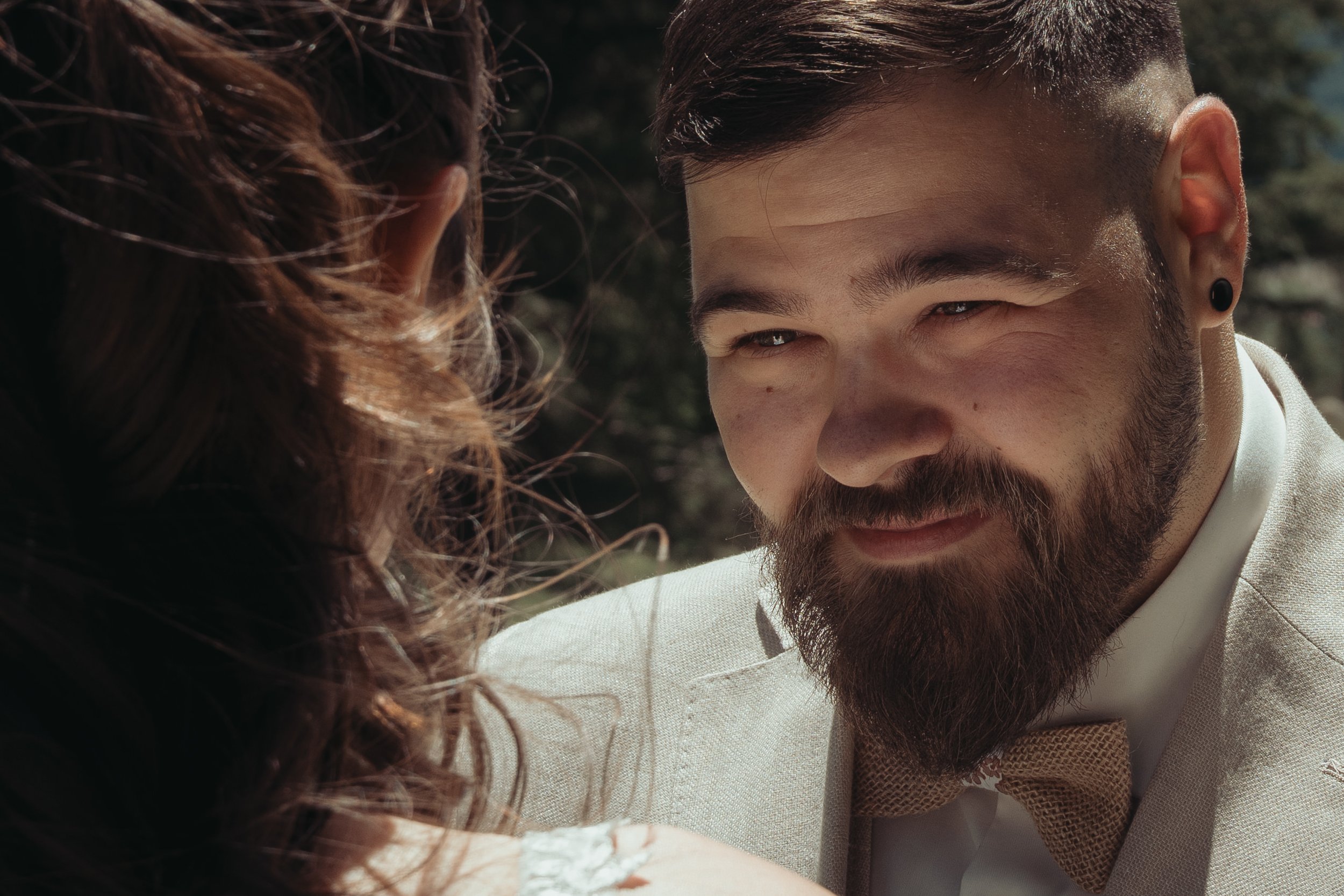 Un homme avec une barbe et une boucle d'oreille noire sourit, vu en gros plan, avec une femme à cheveux bouclés à côté, dans un environnement naturel.