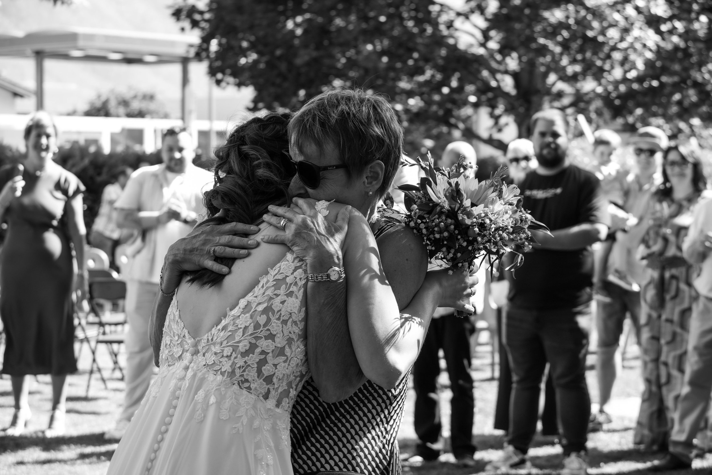 Deux femmes s'embrassent lors d'un moment émouvant, entourées de plusieurs personnes lors d'une occasion en plein air par une journée ensoleillée.