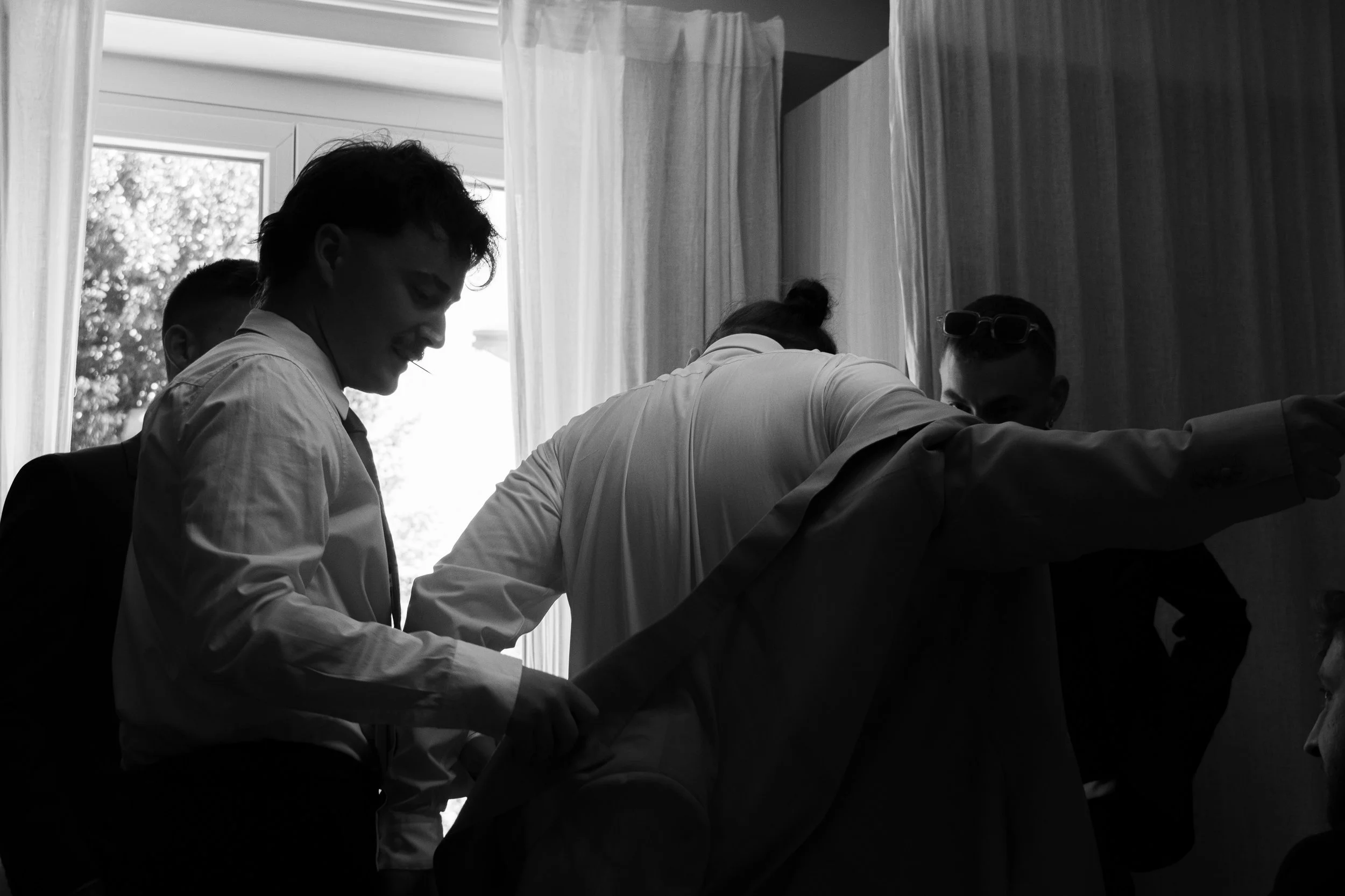 Groupe de jeune hommes habillés en blanc, souriant et interagissant dans une pièce avec des rideaux, en noir et blanc.