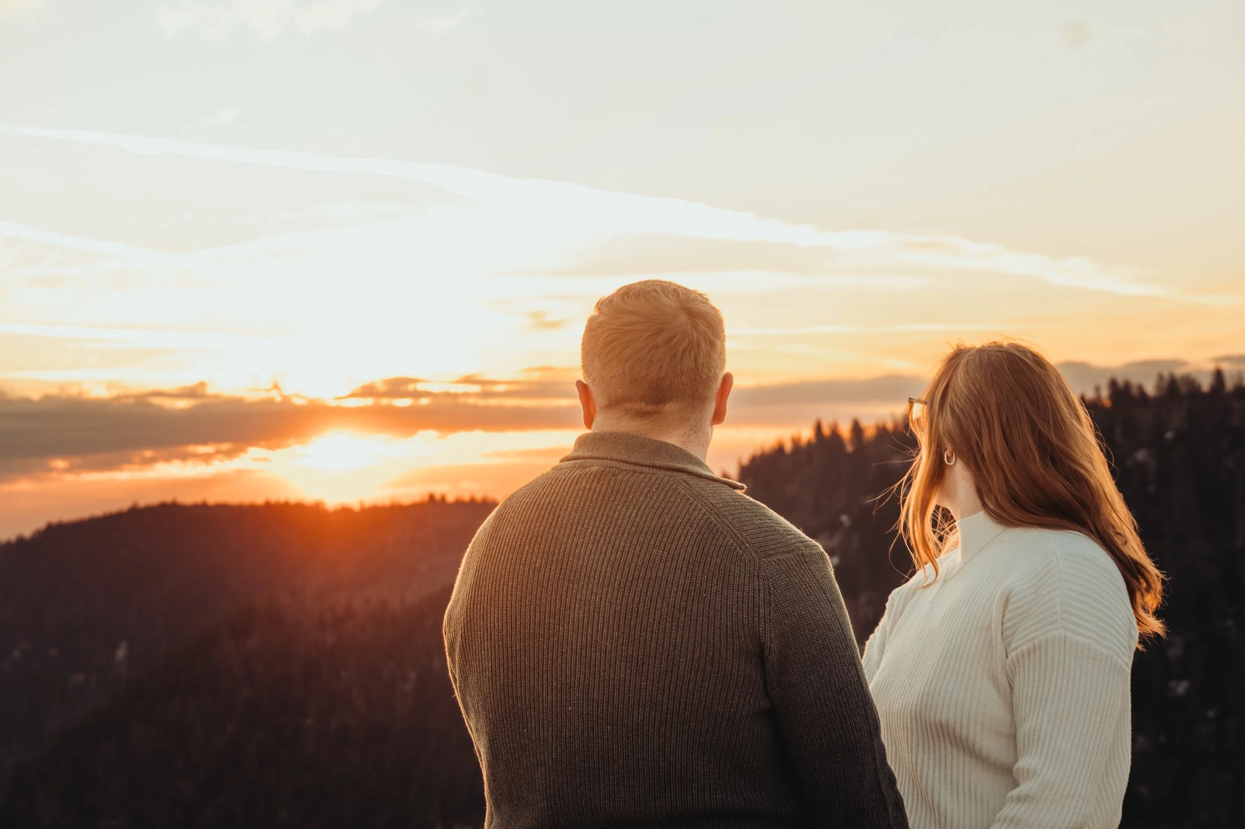 Un homme et une femme regardent un coucher de soleil derrière des collines, créant une scène paisible et romantique.