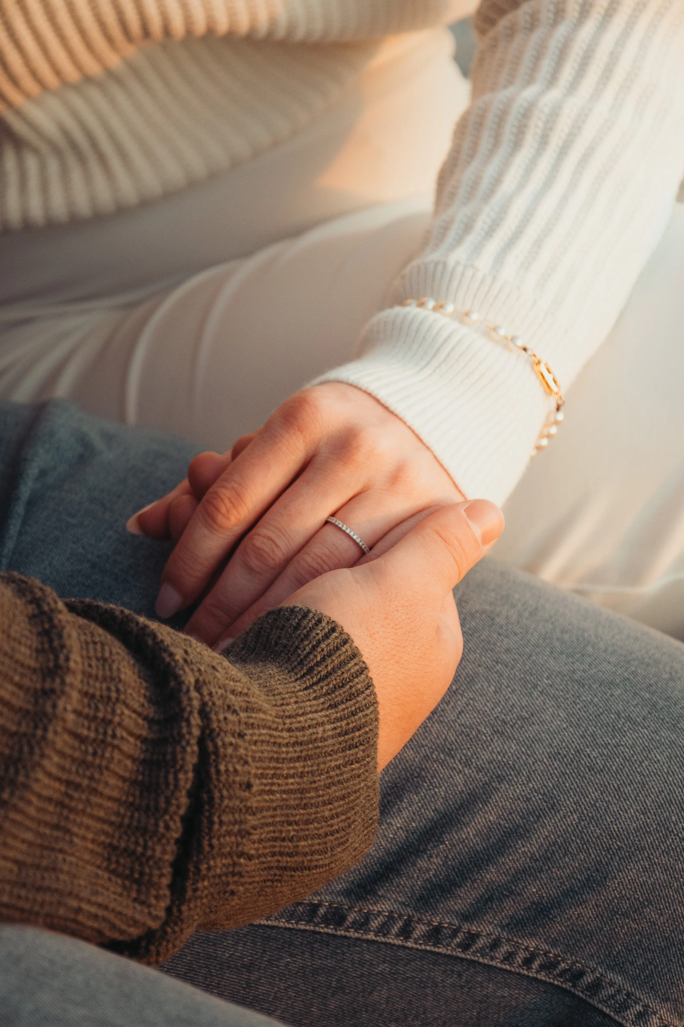 Une personne porte un bracelet en perles dorées et un anneau, tenant la main d'une autre personne. Les deux sont assis côte à côte, portant des vêtements décontractés, avec un fond flou.