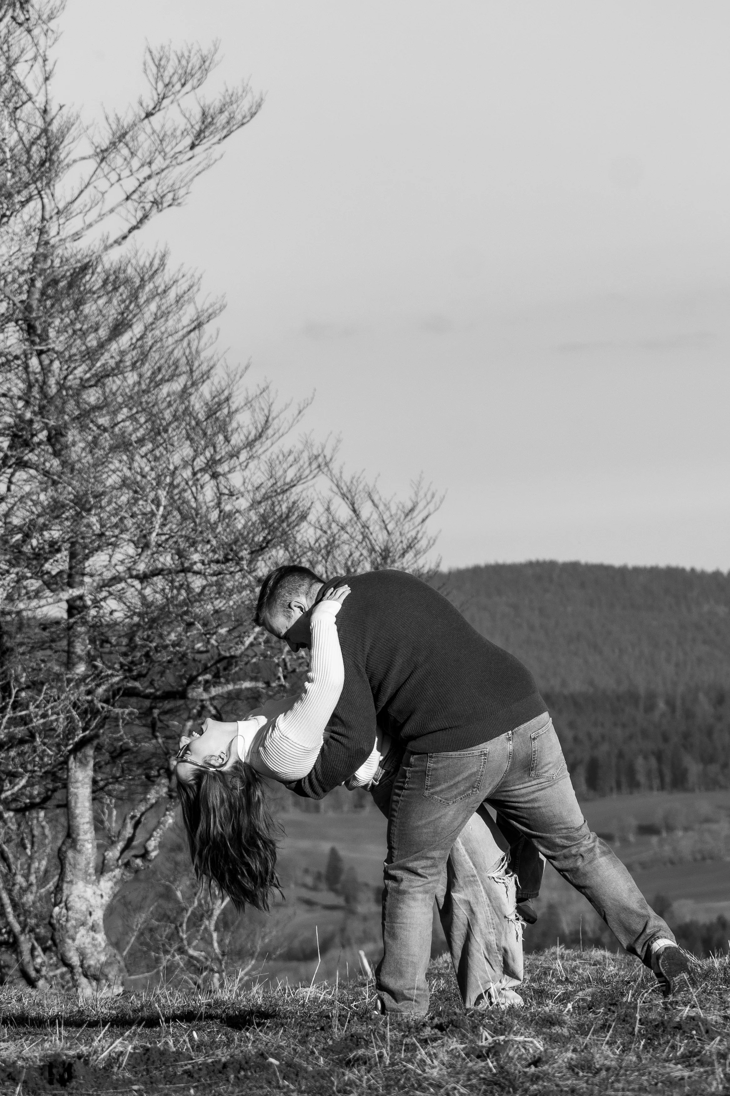 Deux personnes dans un champ en train de danser ou jouer, l'une tenant l'autre en arrière, avec arbres et montagnes en arrière-plan, en noir et blanc.