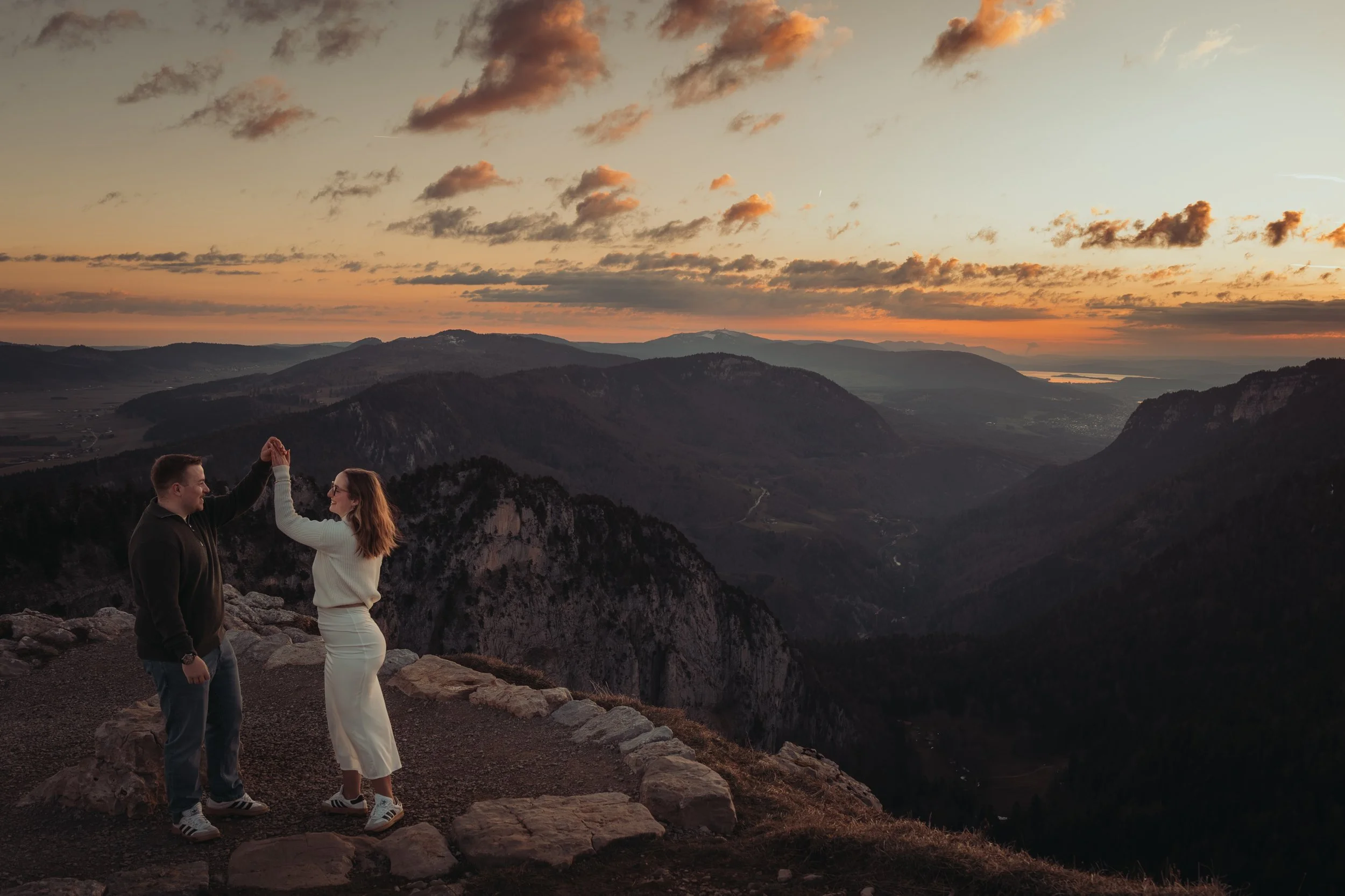 Un homme et une femme se tiennent la main sur un sommet de montagne lors d'un coucher de soleil, avec un paysage vallonné et un ciel nuageux orange en arrière-plan.