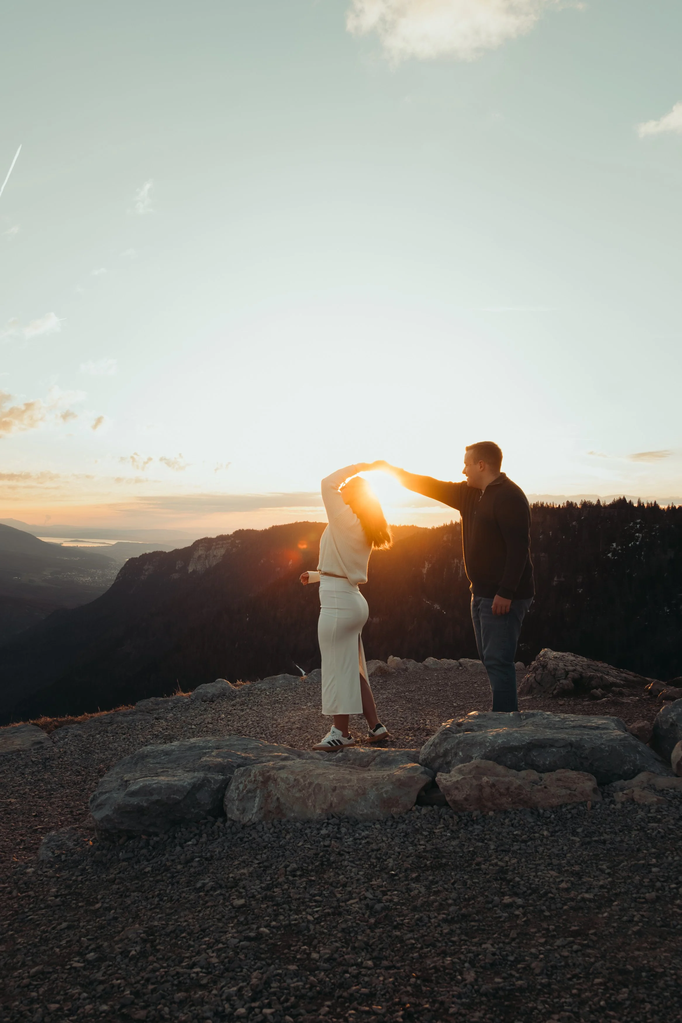 Un couple se tient en face l'un à l'autre sur un rocher lors d'un coucher de soleil avec un paysage de montagnes et de rivières à l'arrière-plan.