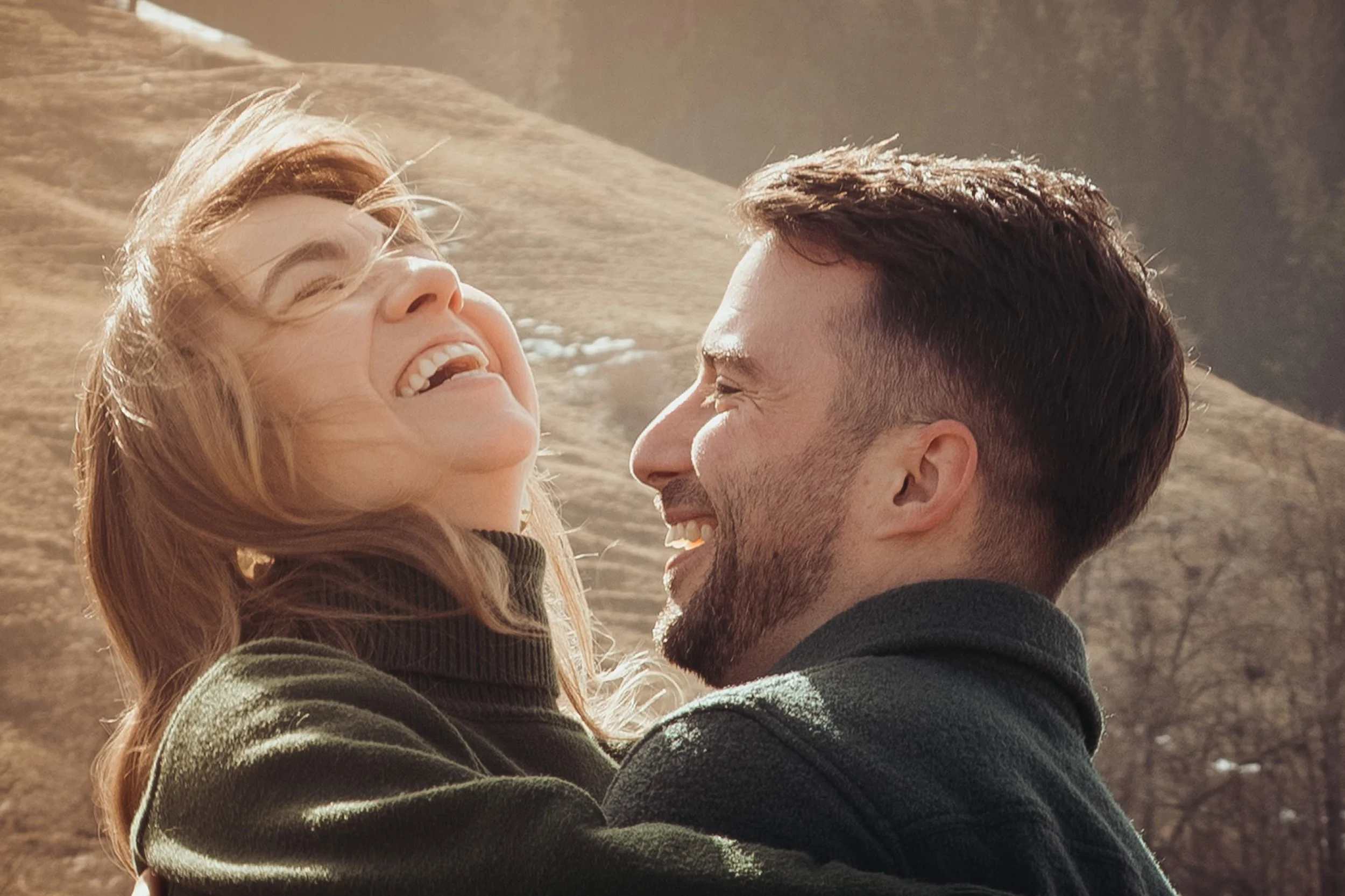 Un couple souriant, adjacent face à face, à l'extérieur avec une lumière douce, en train de rire ou de partager un moment heureux.