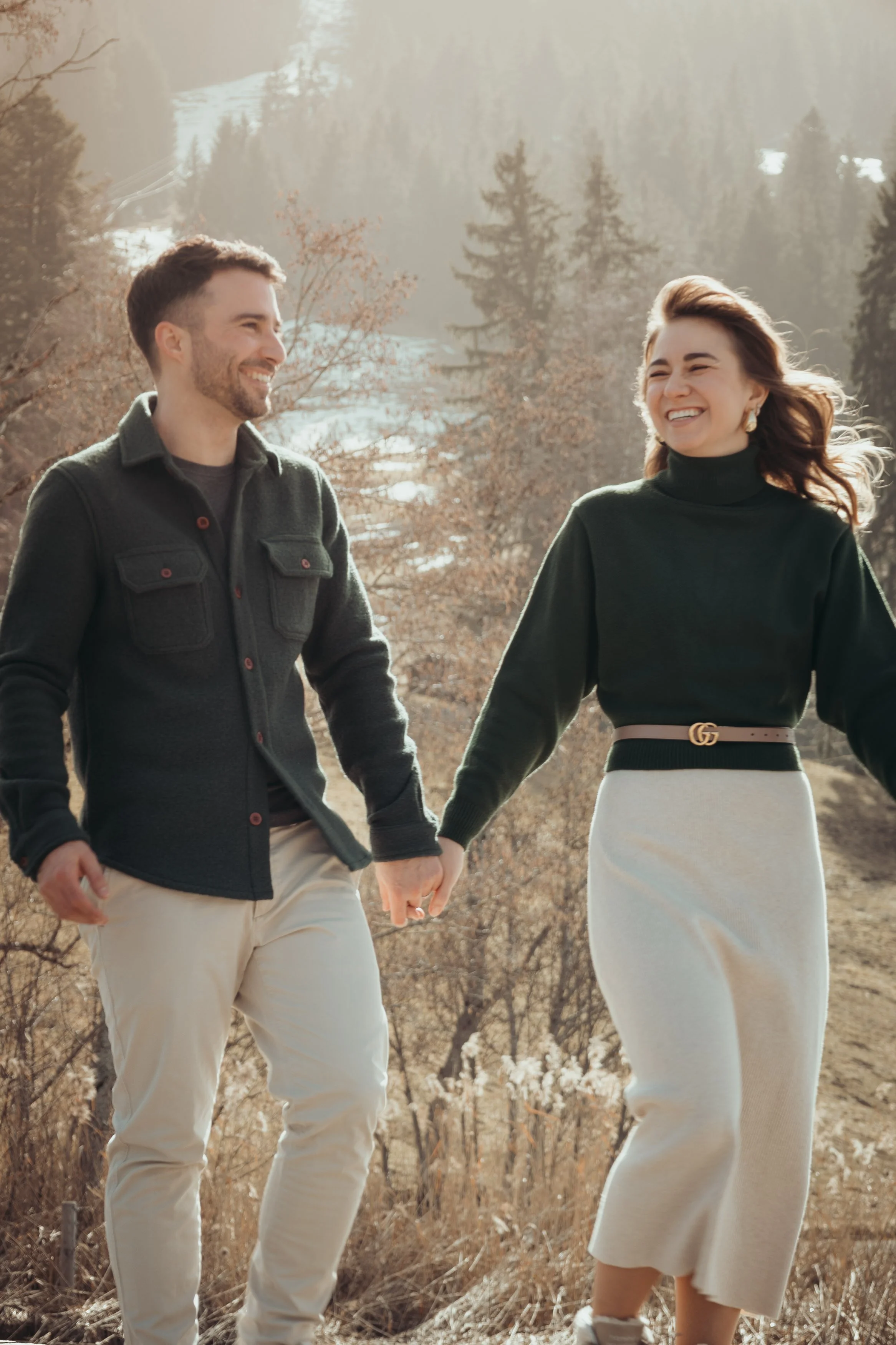 Un couple main dans la main se promenant dans un paysage montagneux, en arrière-plan des arbres et des montagnes enneigées.