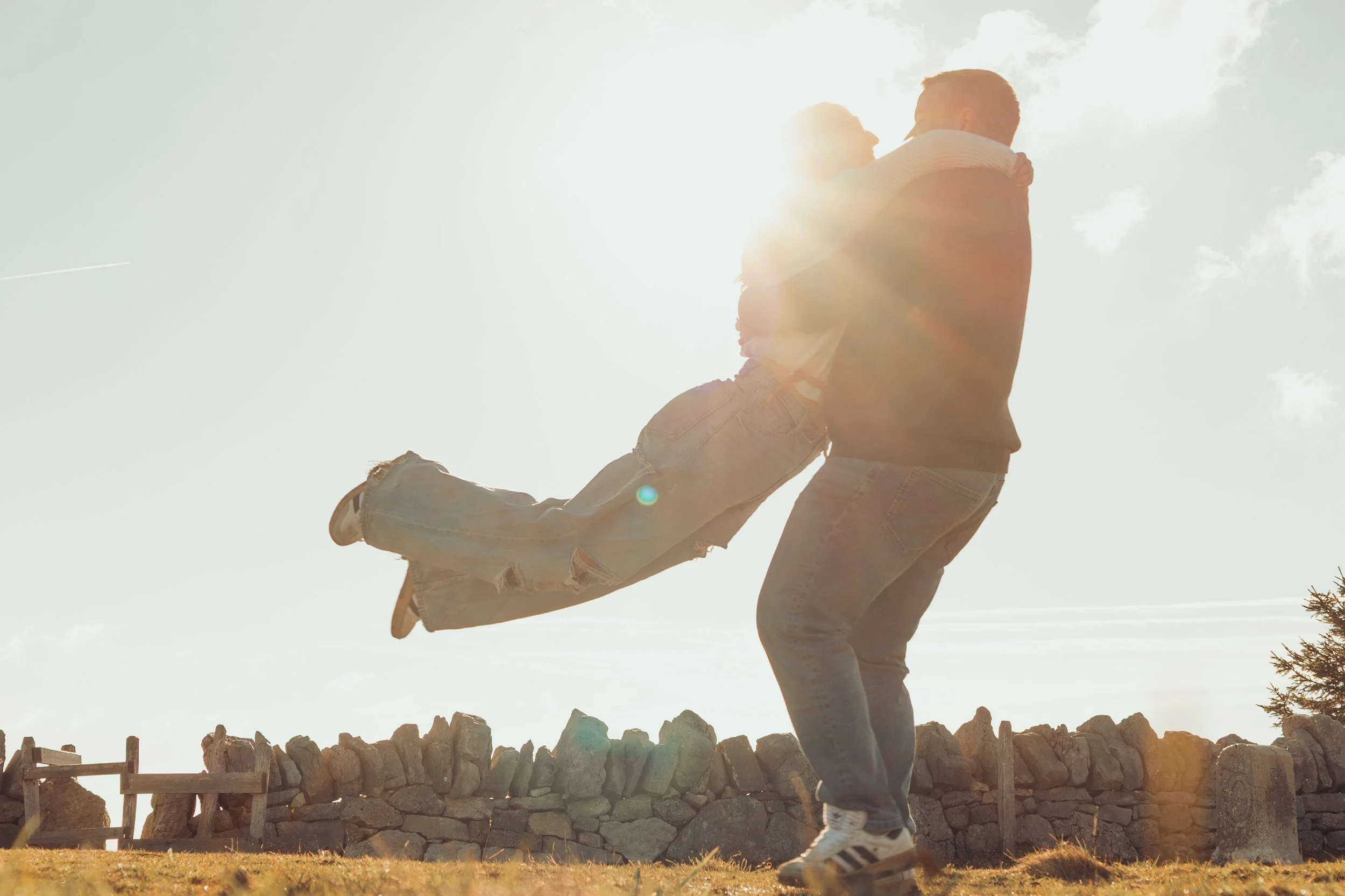 Un homme porte une femme dans ses bras, en plein air au soleil, avec une clôture en pierre derrière eux, donnant une impression de bonheur et d'amour.