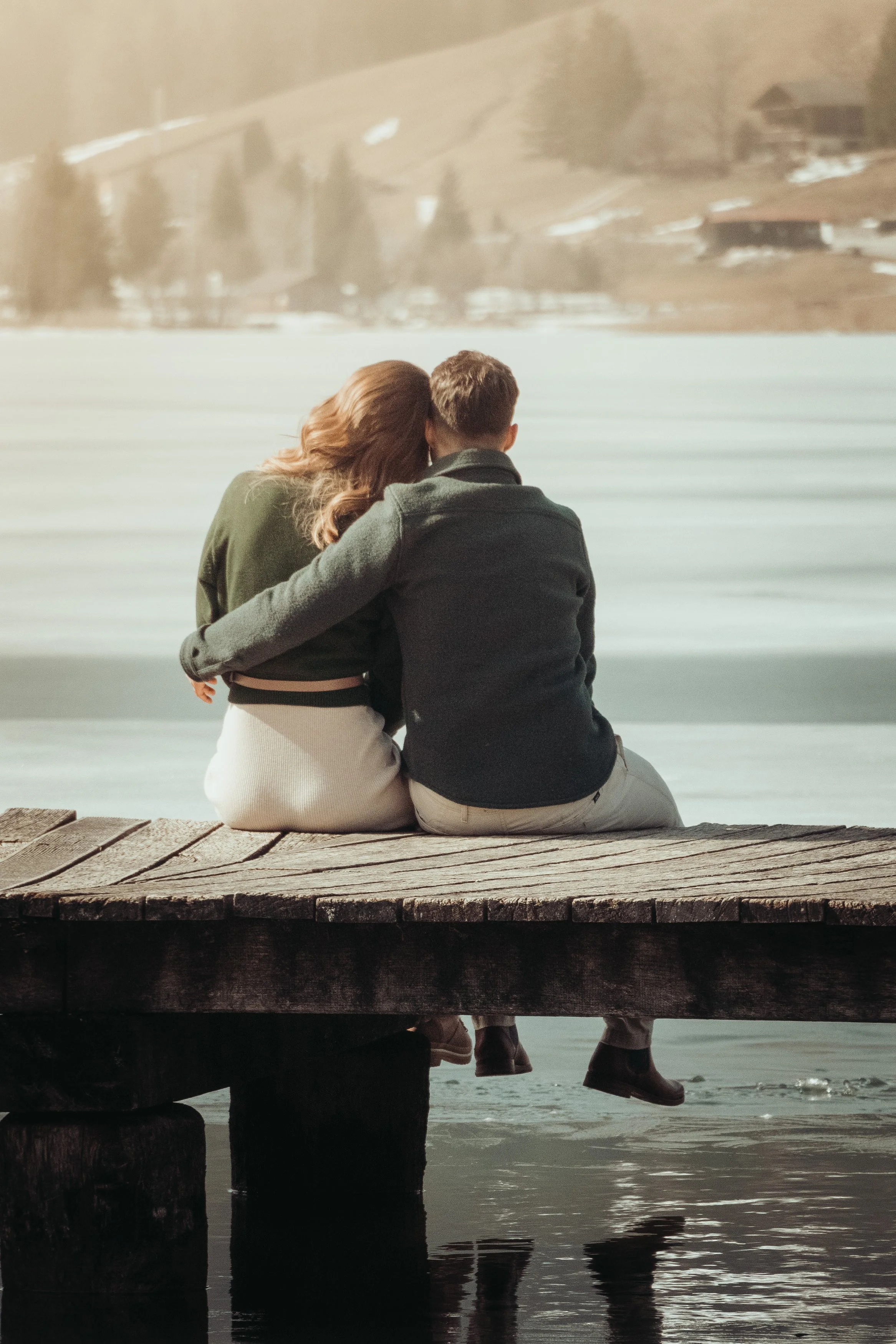 Un couple assis sur un pont en bois, regardant l'eau et un paysage montagnard en arrière-plan. La femme a les cheveux roux, portant un manteau vert et une jupe claire, tandis que l'homme porte un manteau foncé et des chaussures en cuir.