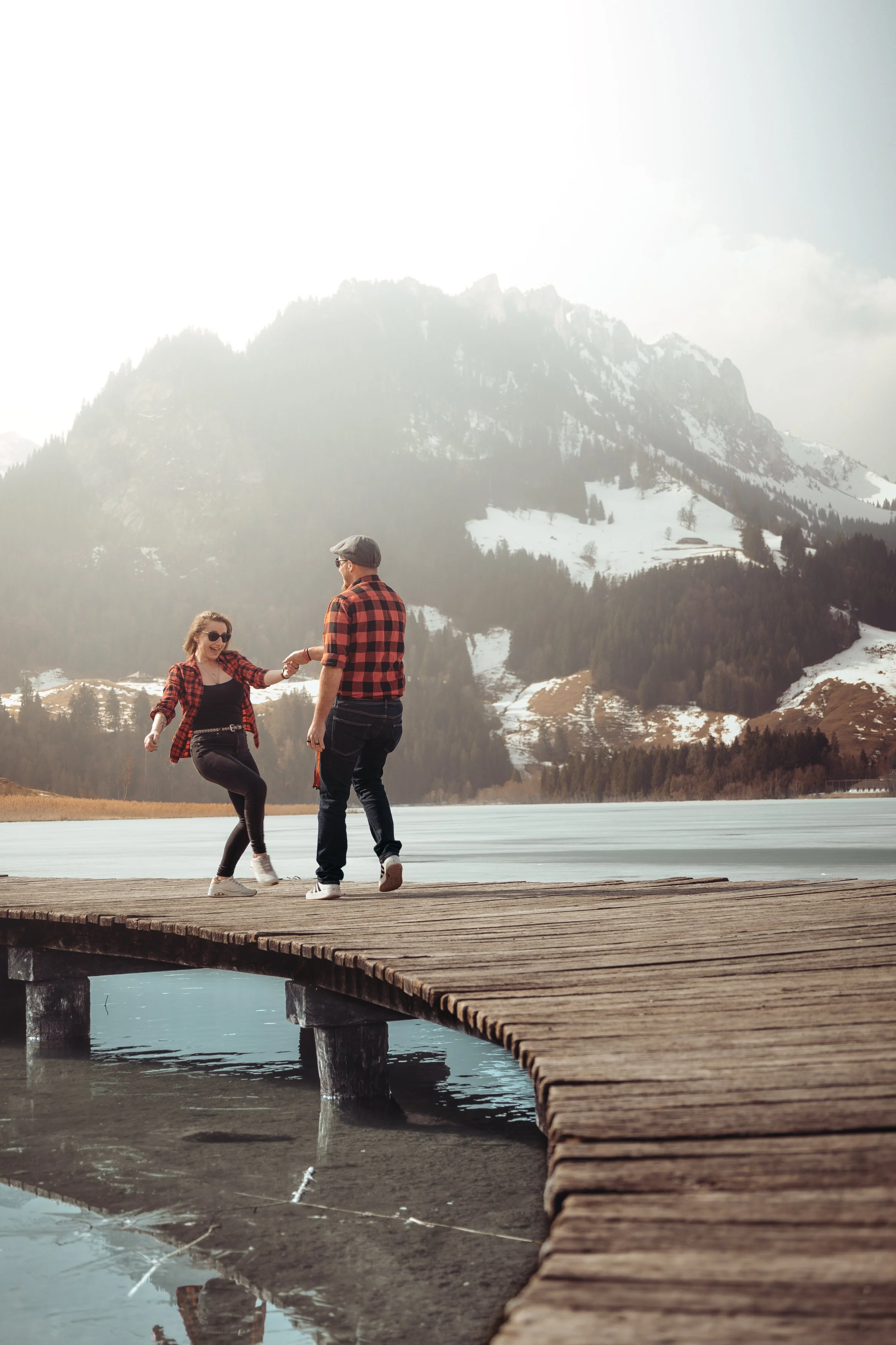 Deux personnes marchent et dansent sur un pont en bois au bord d’un lac avec des montagnes enneigées en arrière-plan.