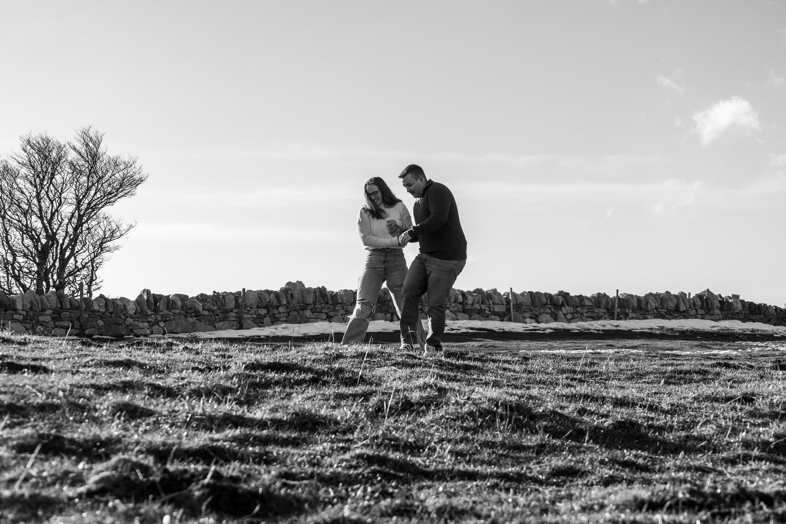 Un couple souriant marche main dans la main dans un champ, avec un mur en pierre en arrière-plan, en noir et blanc.