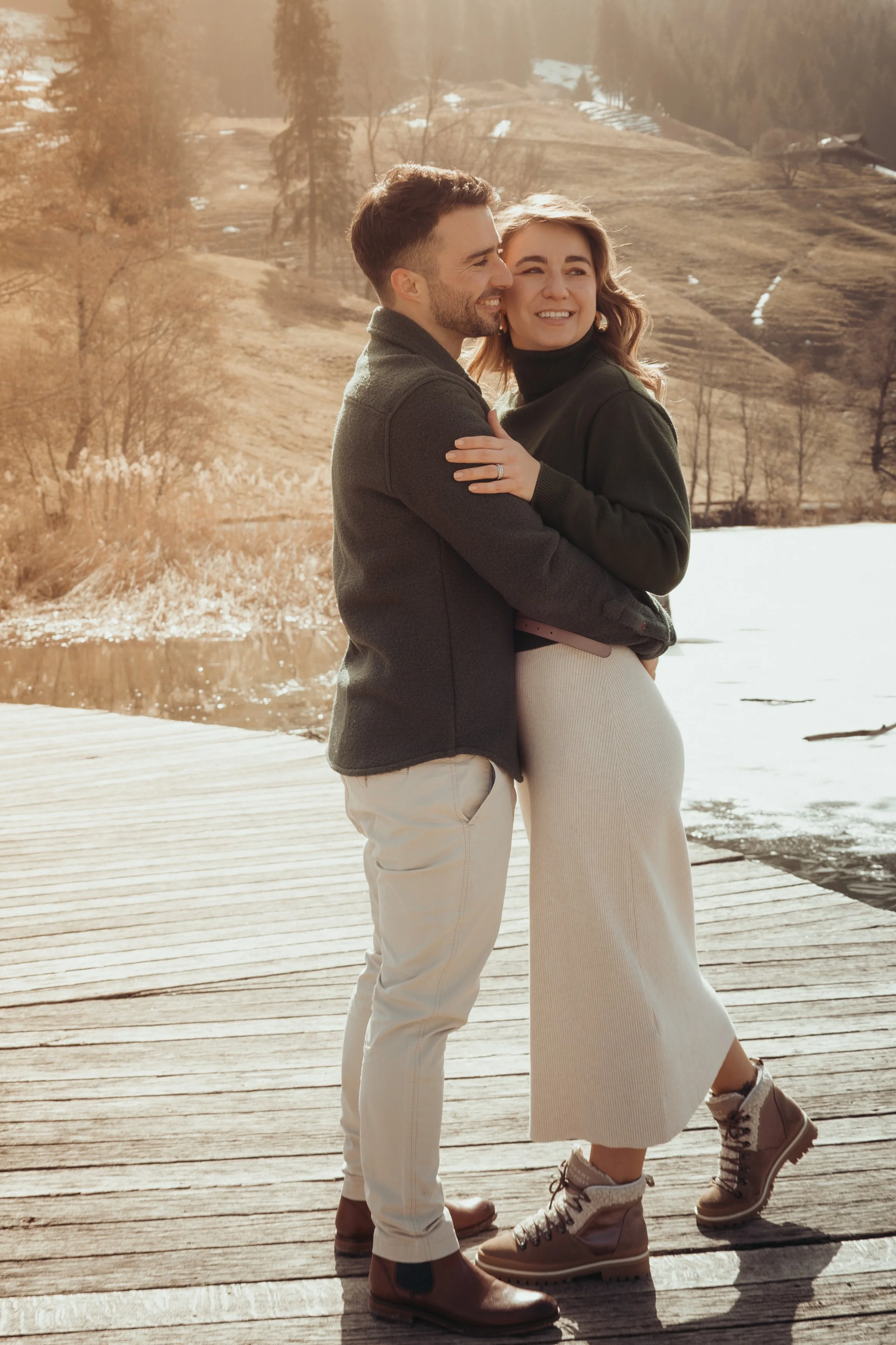 Un couple souriant se tient sur un quai en bois près d'un lac, en arrière-plan des collines et des arbres, avec un éclairage chaud de soleil couchant.