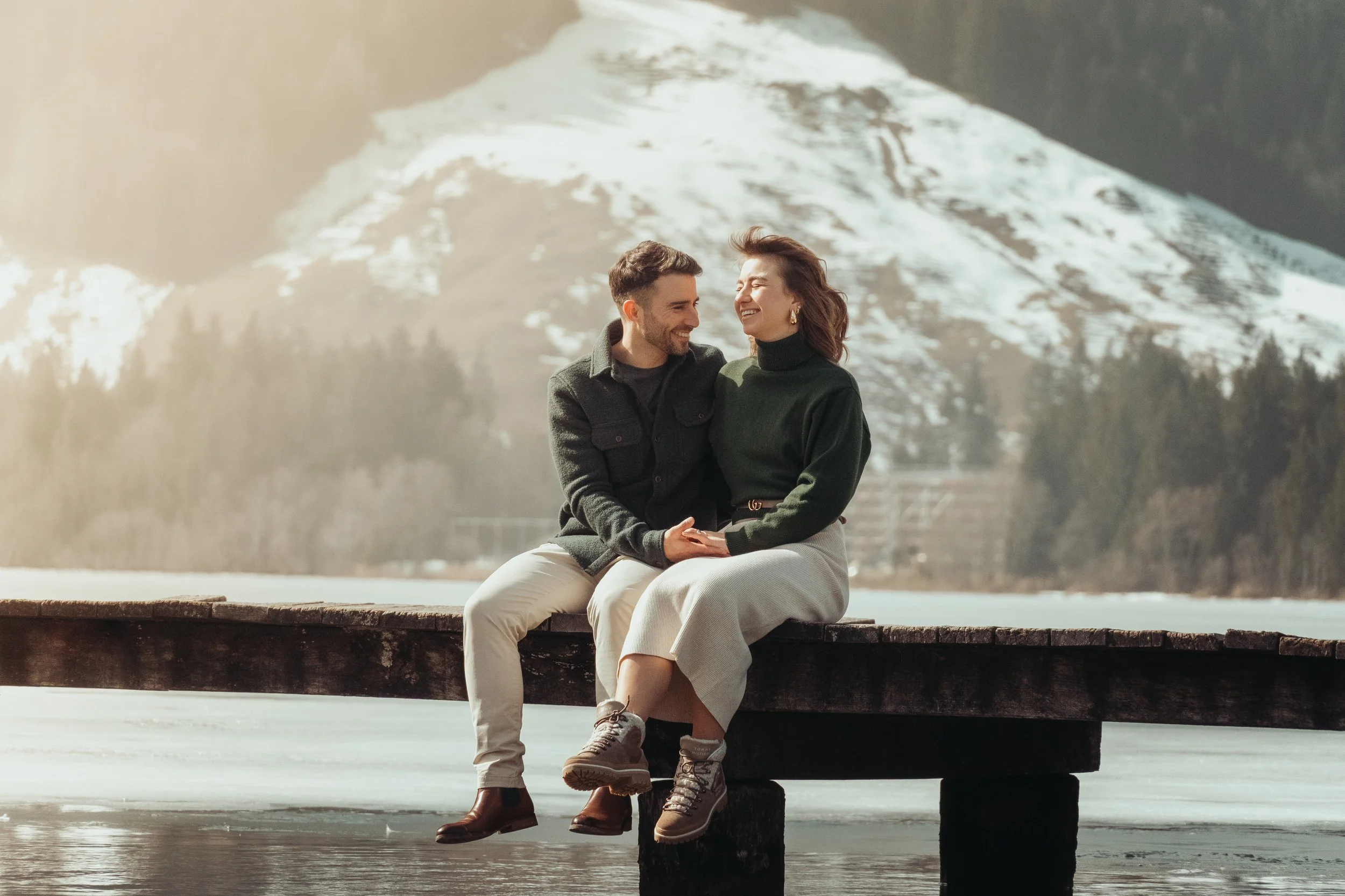 Un couple souriant et riant, assis sur un quai en bois près d'un lac, avec des montagnes enneigées en arrière-plan, en hiver.