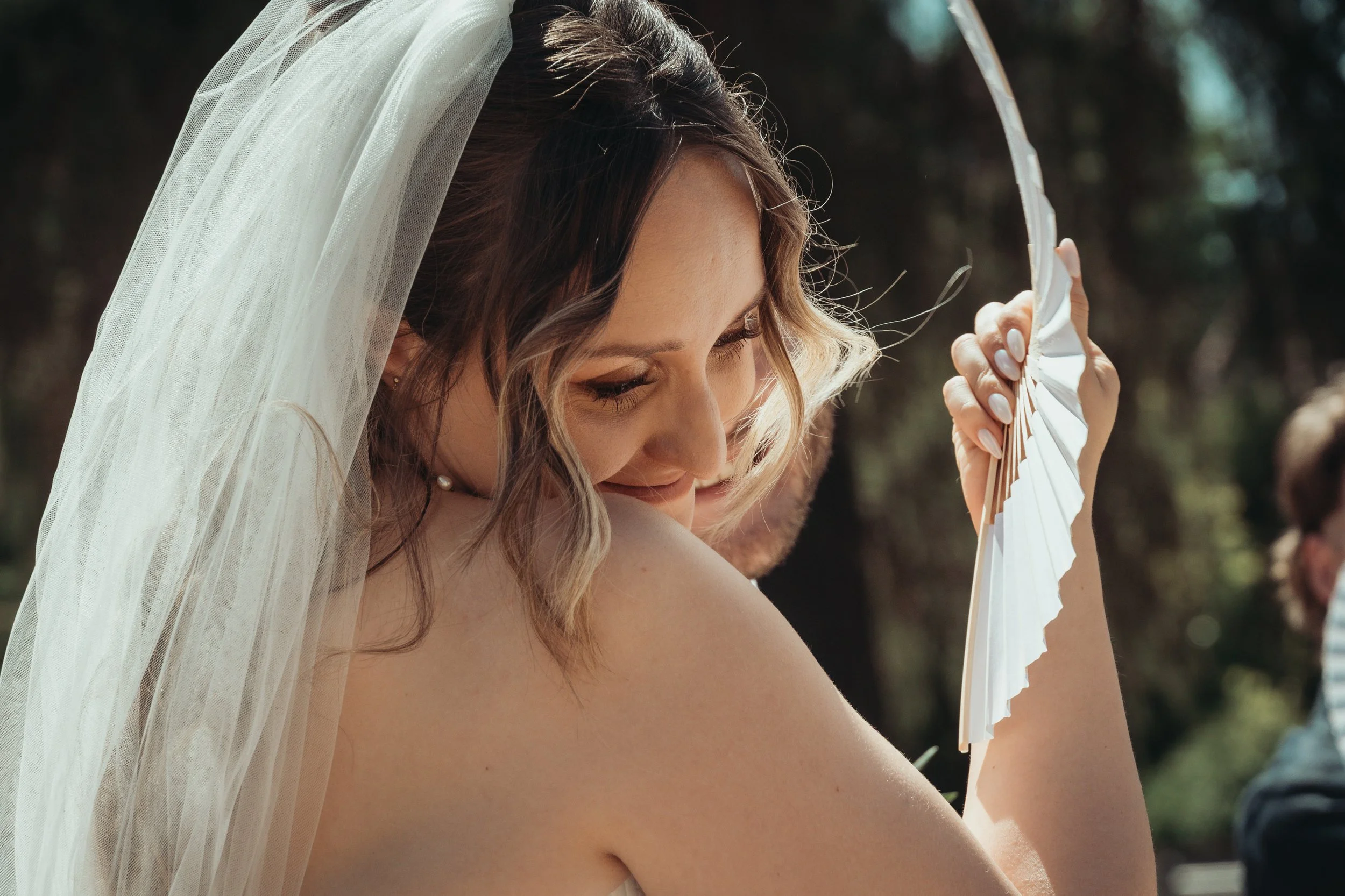 Une femme portant un voile blanc et une robe de mariage, tenant un éventail blanc en tissu, dans un environnement extérieur ensoleillé.