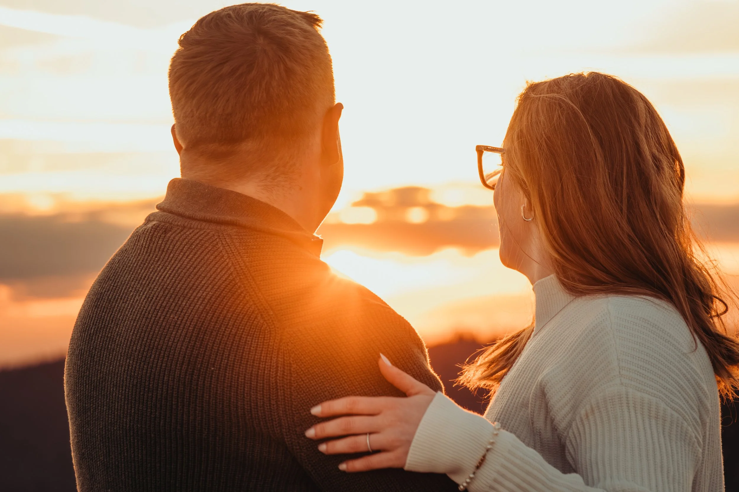 Un couple regardant le coucher du soleil, lui portant une veste sombre et elle portant des lunettes, s'enlaçant tendrement.