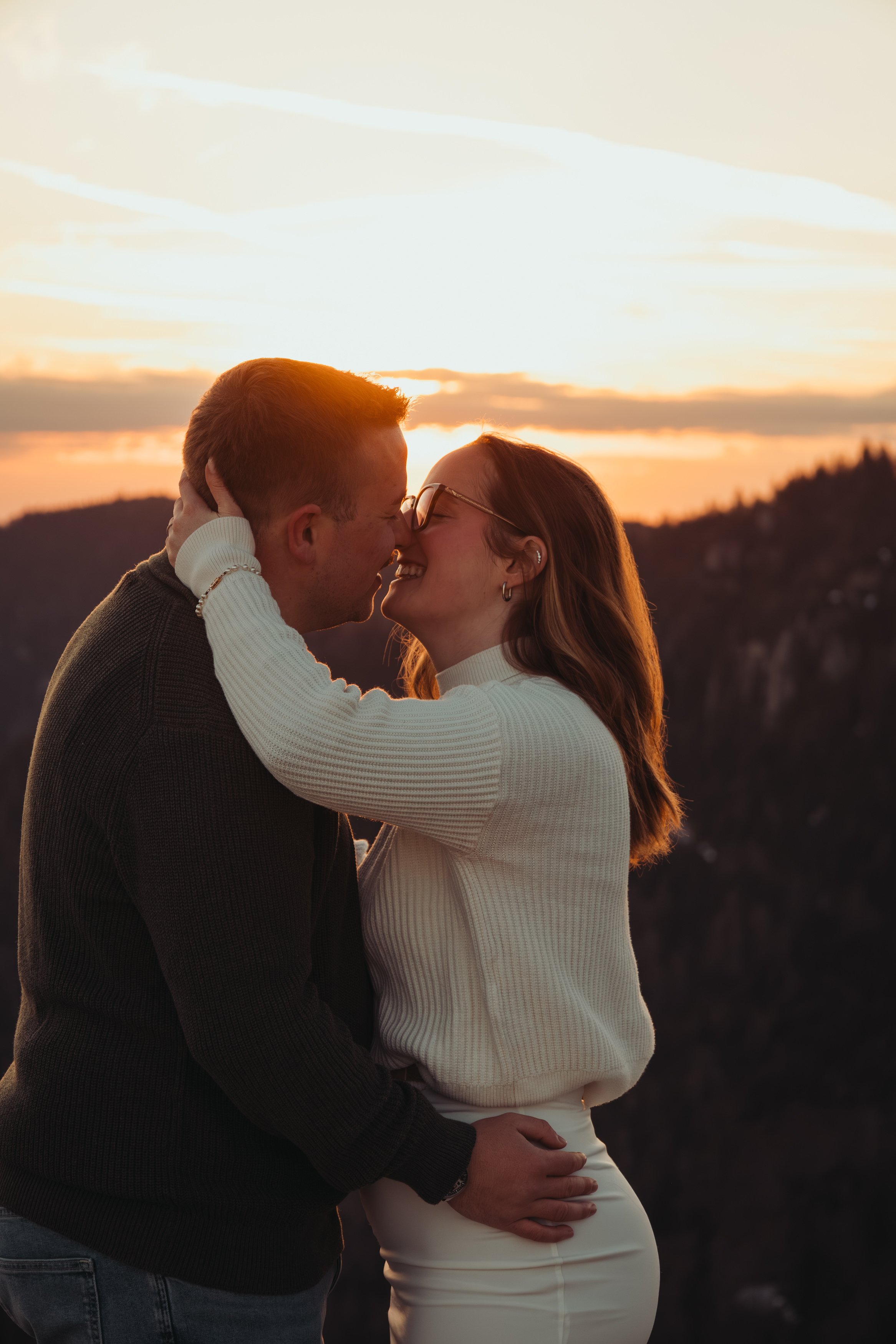 Un couple s'embrasse tendrement lors d'un coucher de soleil, avec un paysage montagneux en arrière-plan.