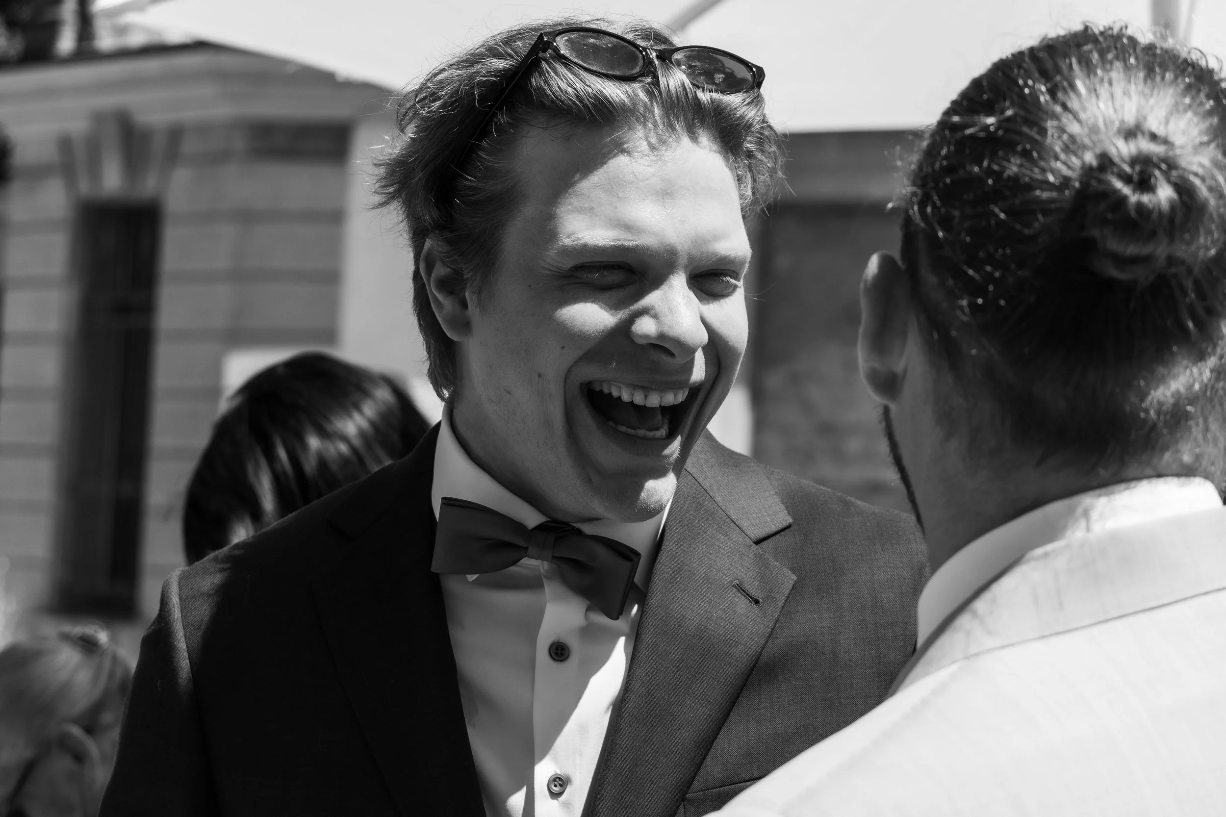 Homme souriant en costume noir avec une cravate, discutant avec une femme à une cérémonie extérieure.