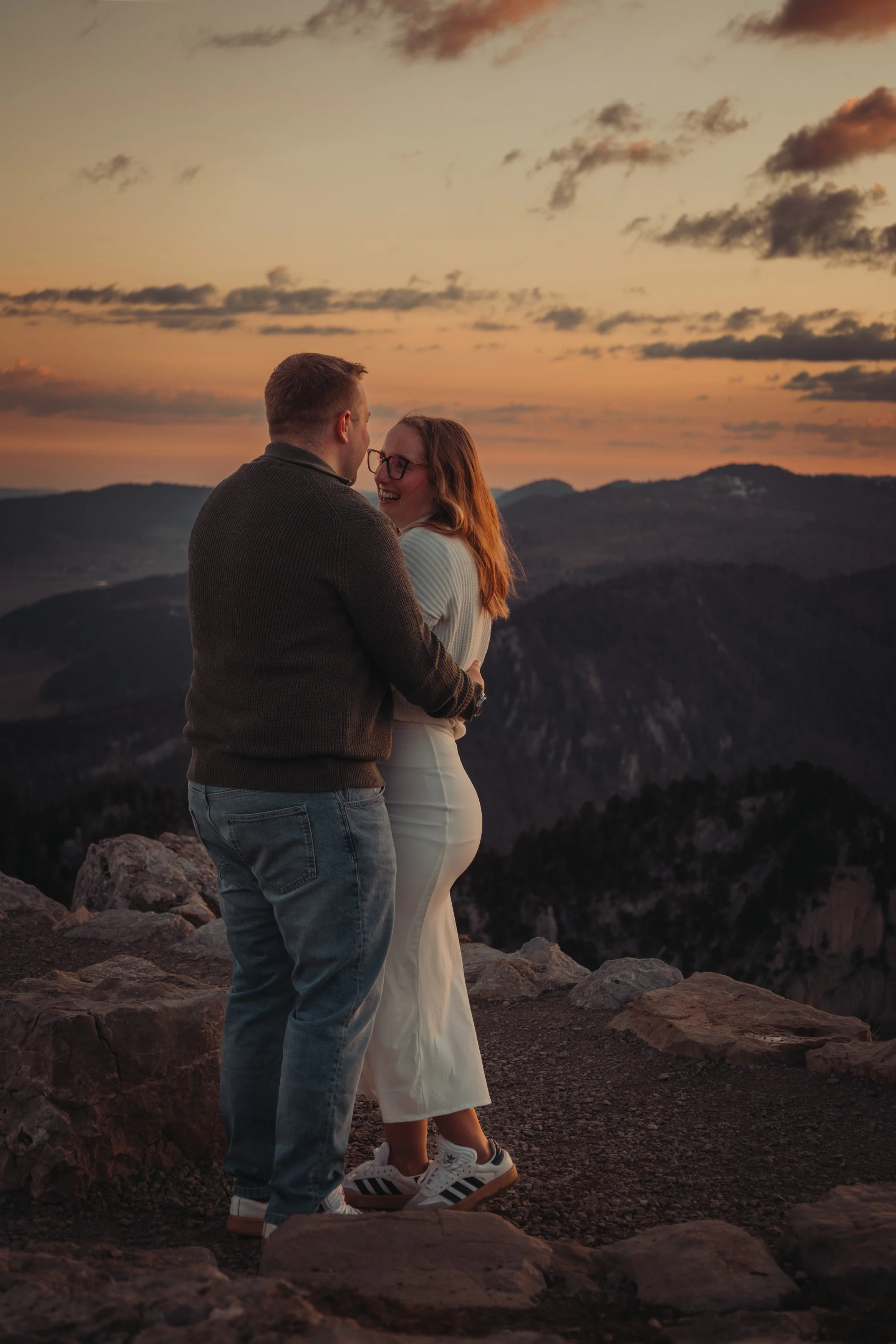 Un couple souriant se tient proche l'un de l'autre sur un sommet rocheux lors d'un coucher de soleil, avec un paysage de montagnes et un ciel coloré en arrière-plan.