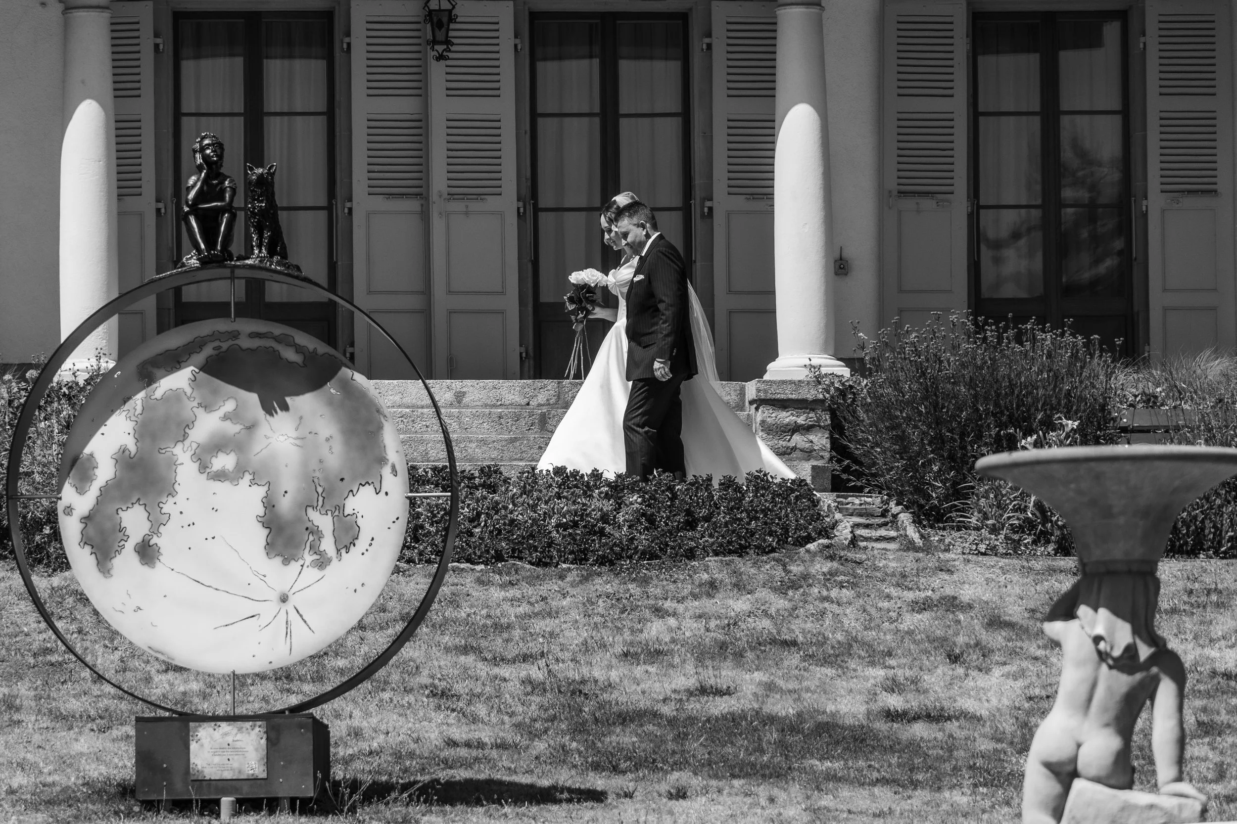 Une scène en noir et blanc d'un couple en costume de mariage qui se tient devant une maison avec des volets en bois, un lampadaire suspendu, et une sculpture d'une enfant avec un chien en haut d'une arche. Le couple tient des fleurs, la femme porte u