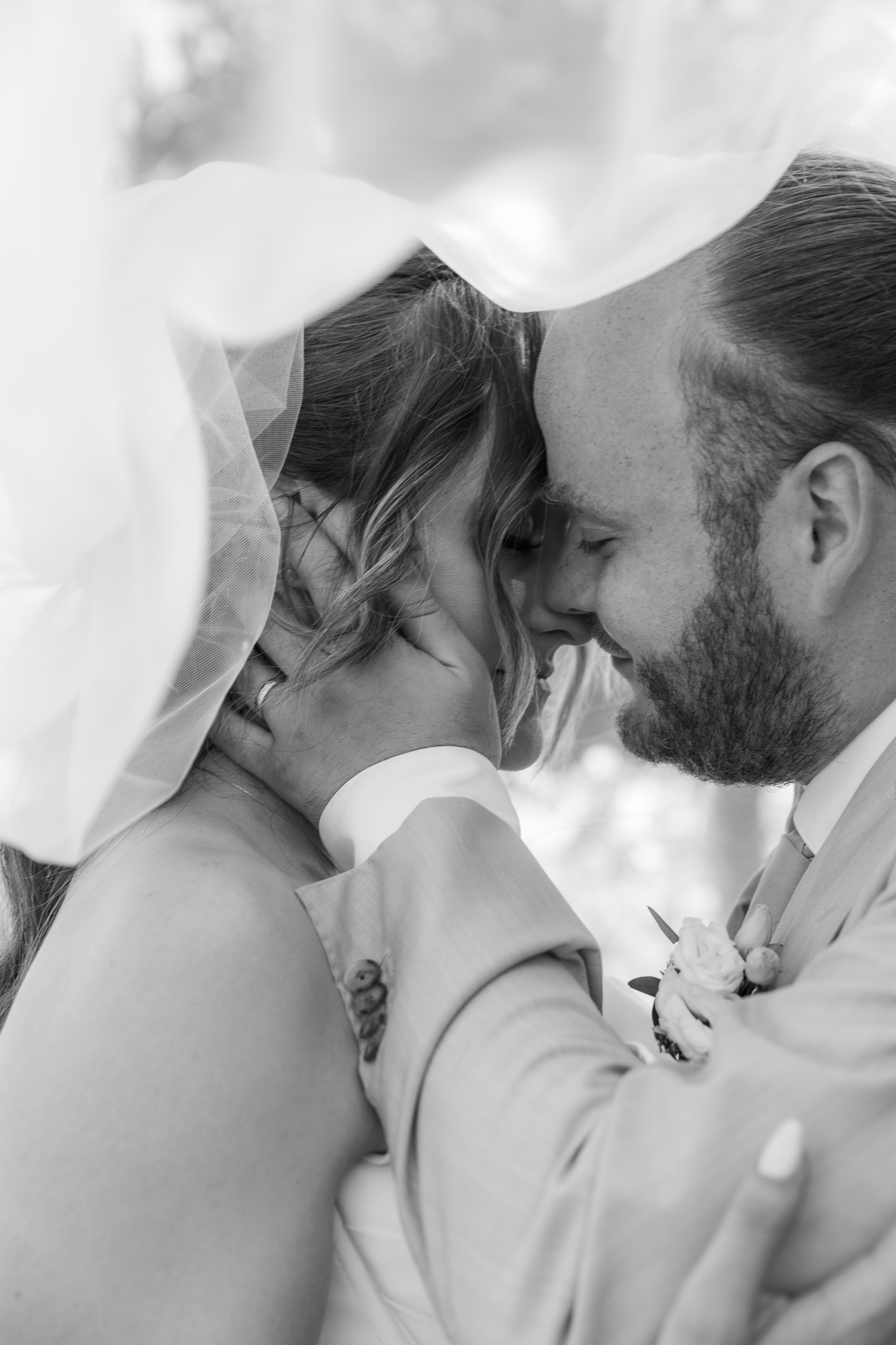 Un couple échange un baiser lors de leur mariage, la femme porte une robe de mariée et une veste, l'homme porte une chemise et a un boutonnière avec des fleurs. L'image est en noir et blanc.