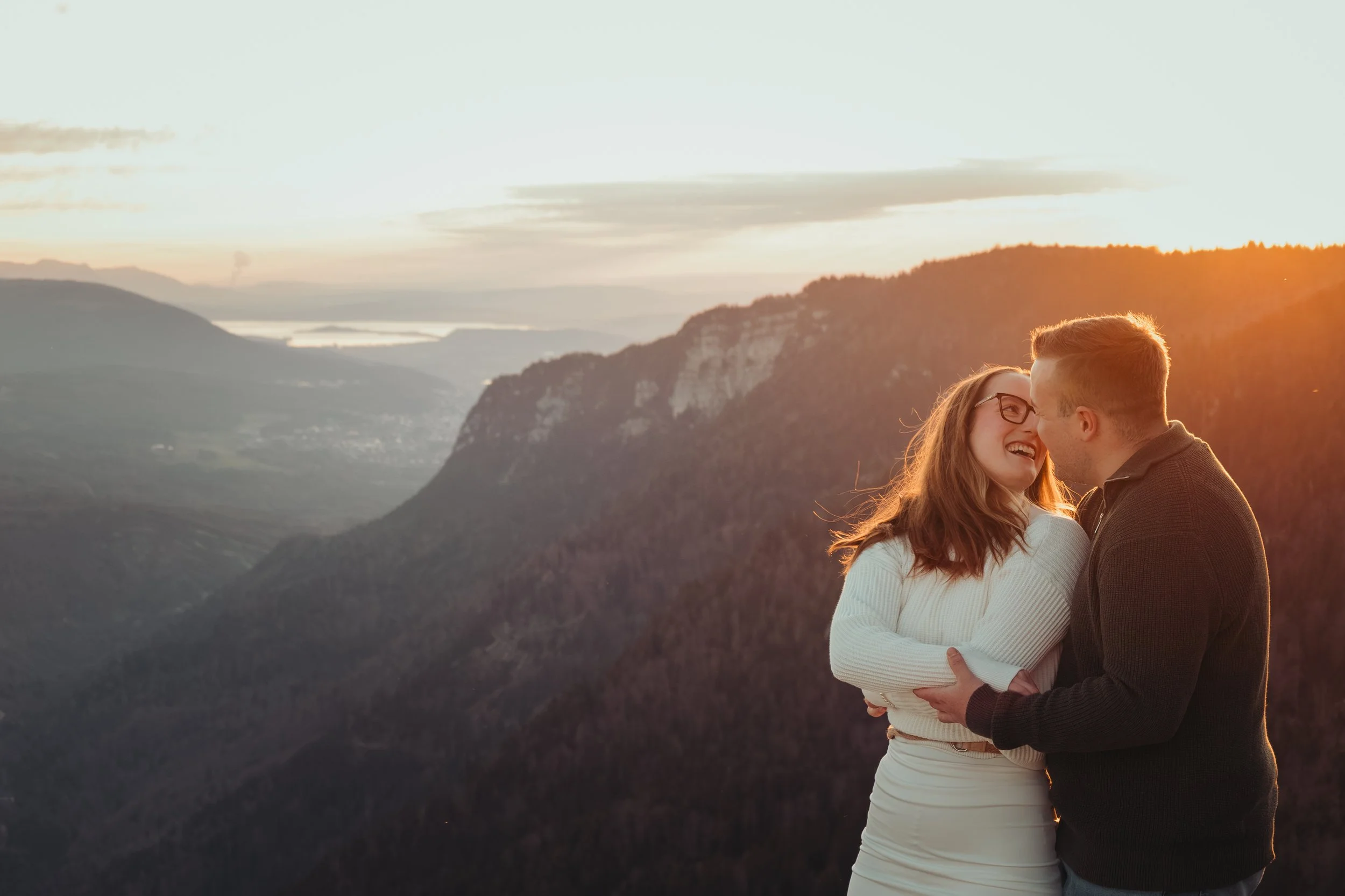 Un couple souriant se tient près d'un paysage de montagnes au coucher du soleil, avec la femme portant des lunettes et le homme embrassant le front de la femme.
