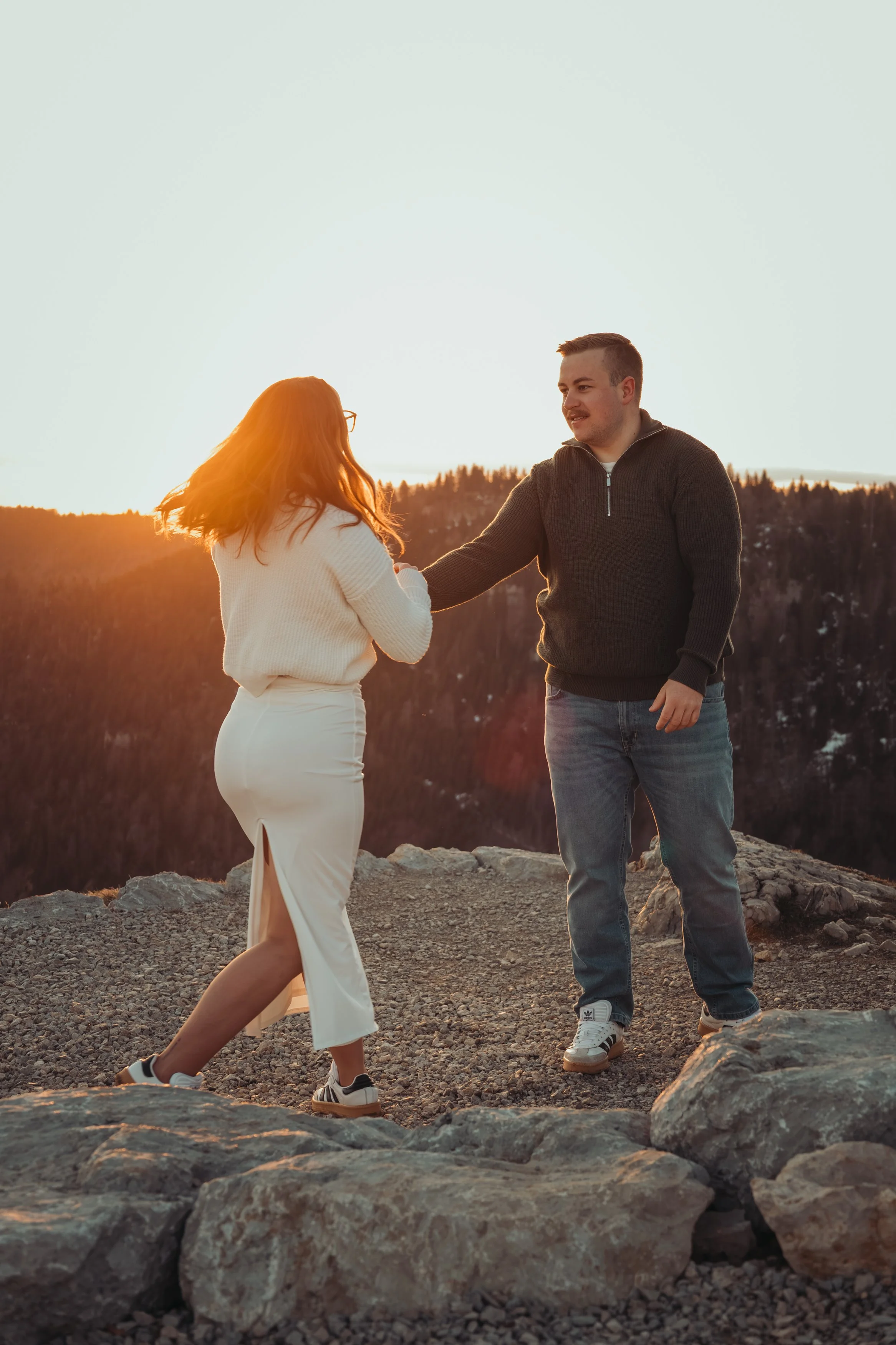 Un homme et une femme se tiennent la main sur un rocher lors d'un coucher de soleil, avec des montagnes en arrière-plan.