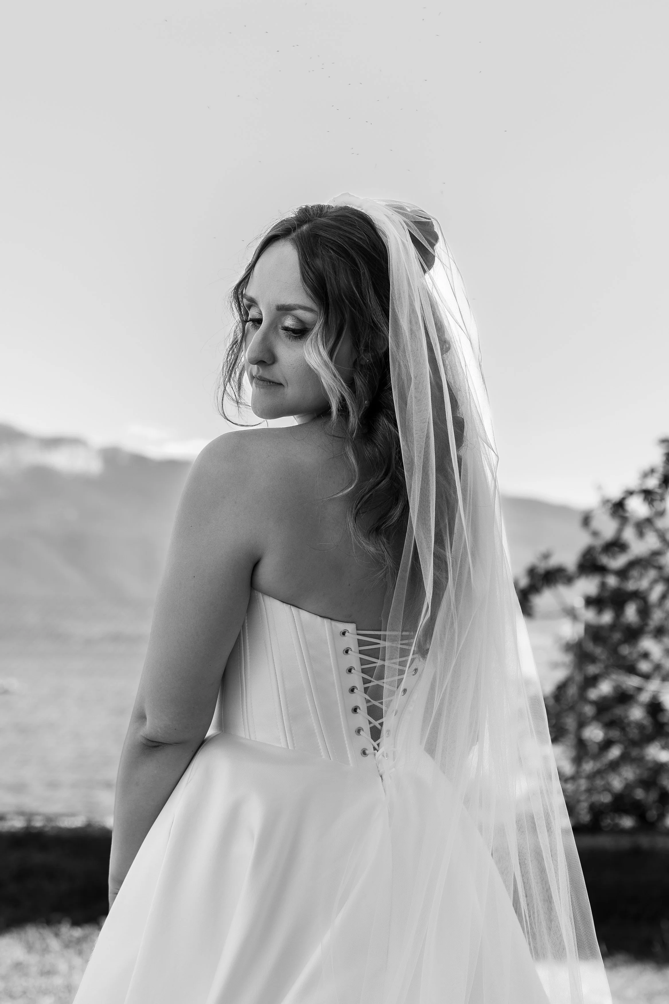 Une femme en robe de mariée avec un voile, vue de profil, dans un paysage naturel en noir et blanc.