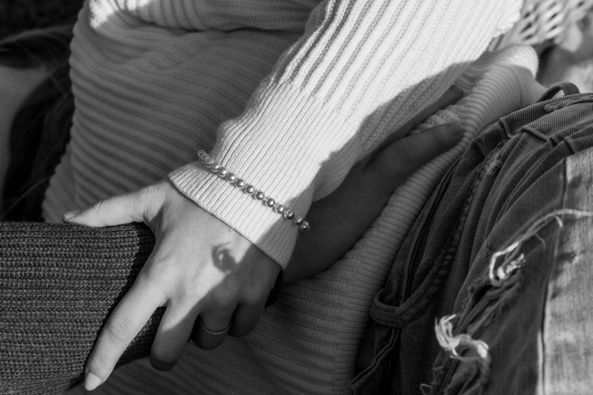 Deux mains entrelacées reposant sur le genou d'une personne, portant des vêtements en laine ou en coton, avec un bracelet de perles sur le poignet. Image en noir et blanc.