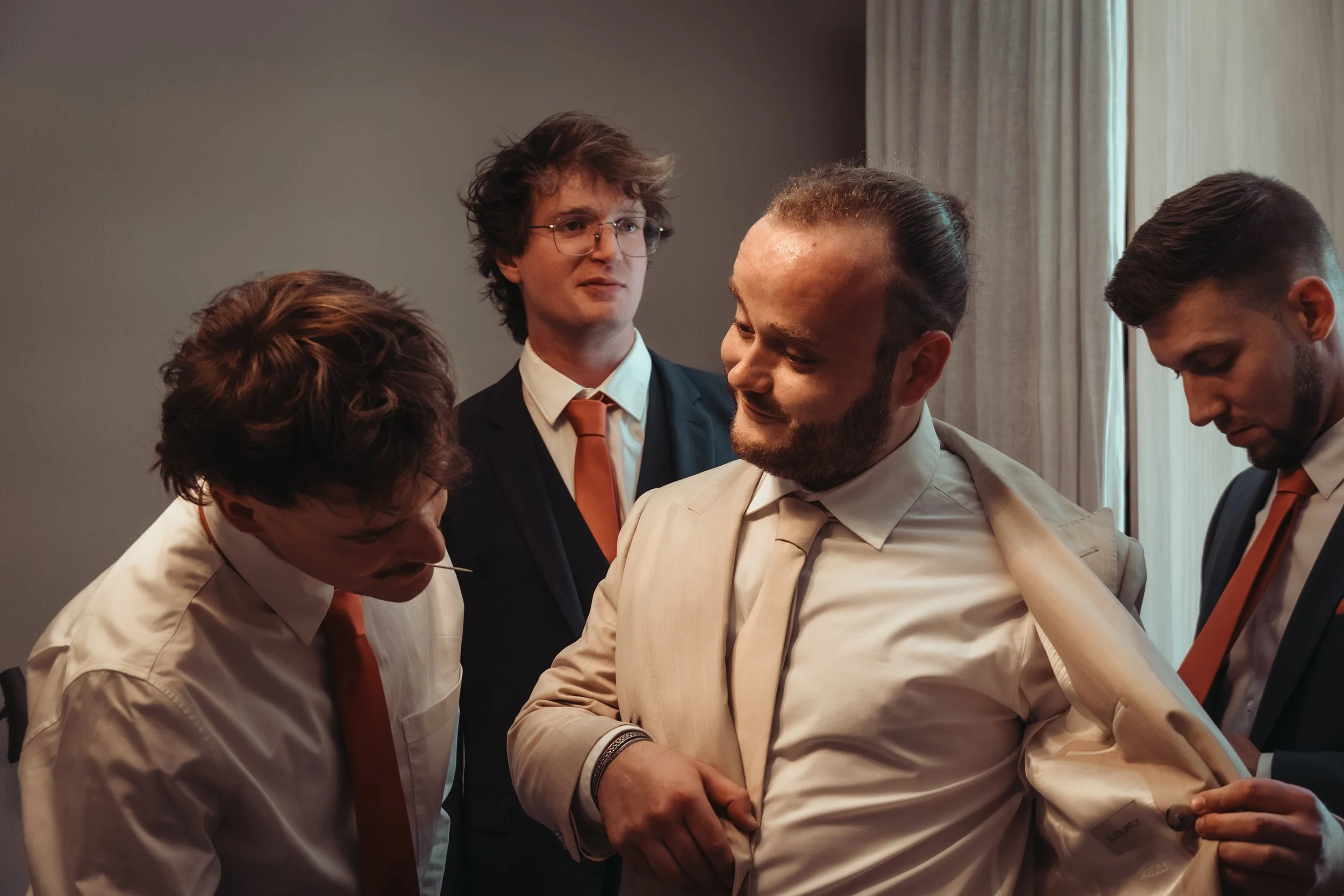 Un groupe de jeunes hommes en costume, certains avec des cravates orange, qui semblent en train de discuter ou de se préparer pour un événement formel dans une pièce avec rideaux pale en arrière-plan.