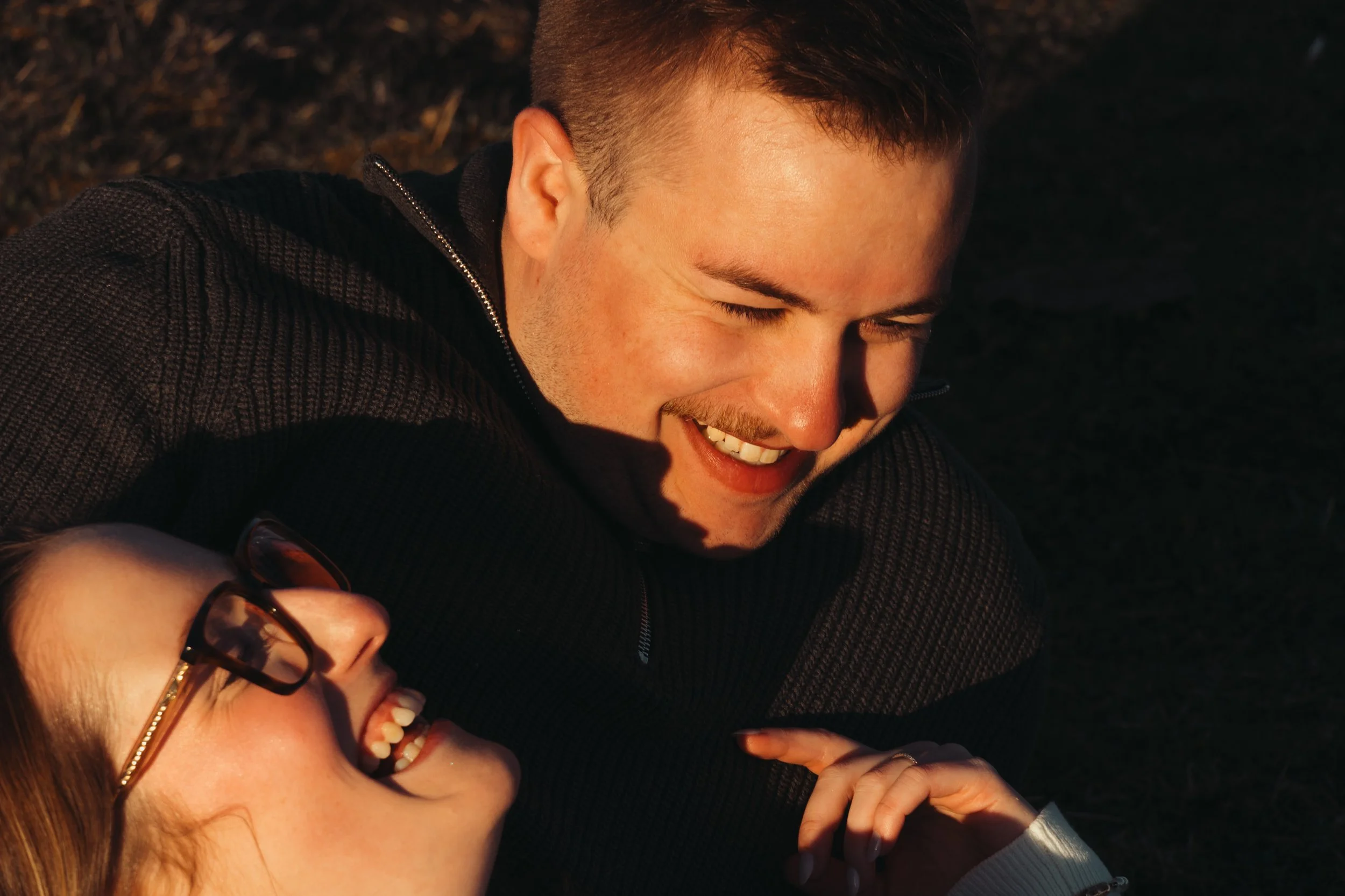 Deux personnes, une femme avec des lunettes et une autre personne dont on voit juste le visage souriant, partagent un moment heureux dehors, probablement en plein air. La femme porte des lunettes à monture noire, semble rire ou sourire, et a les chev