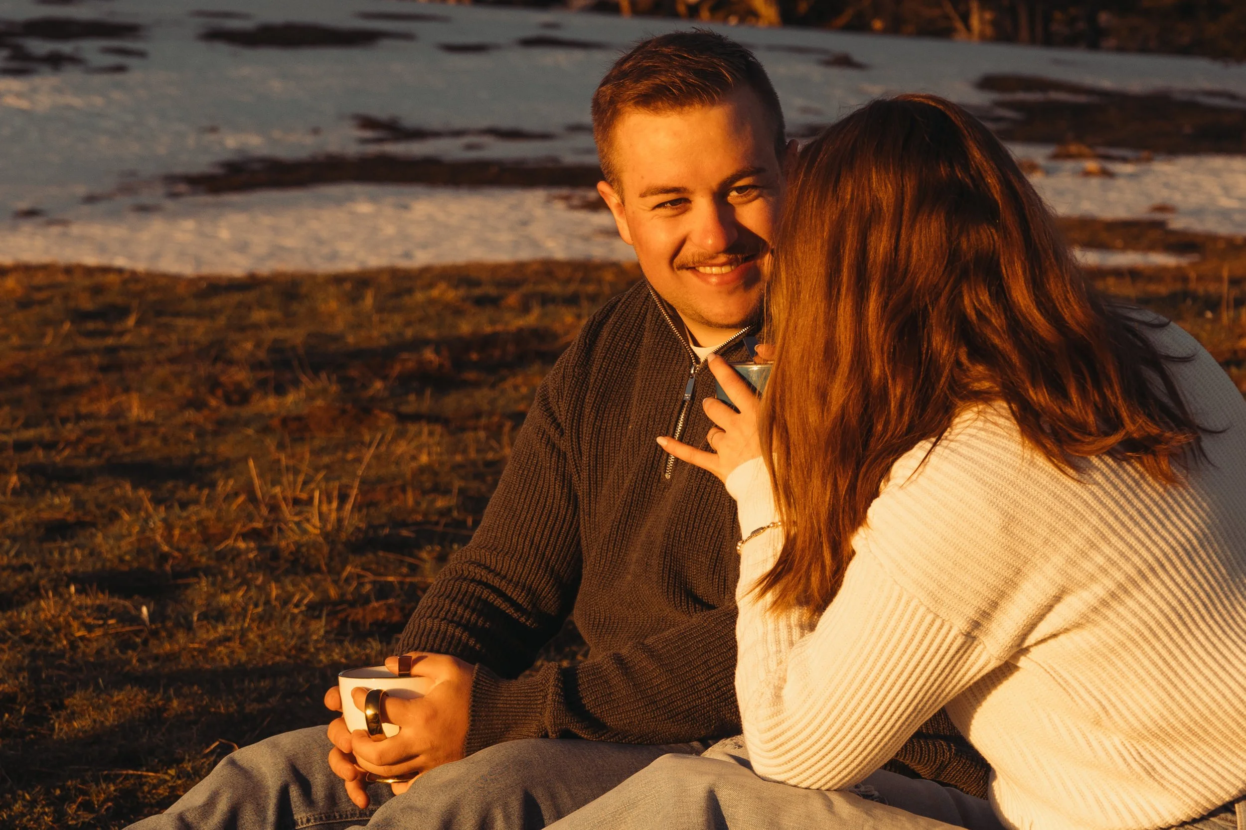 Un homme et une femme assis ensemble près d'un lac au coucher du soleil, ils discutent et semblent heureux. La femme tient une tasse dans ses mains.