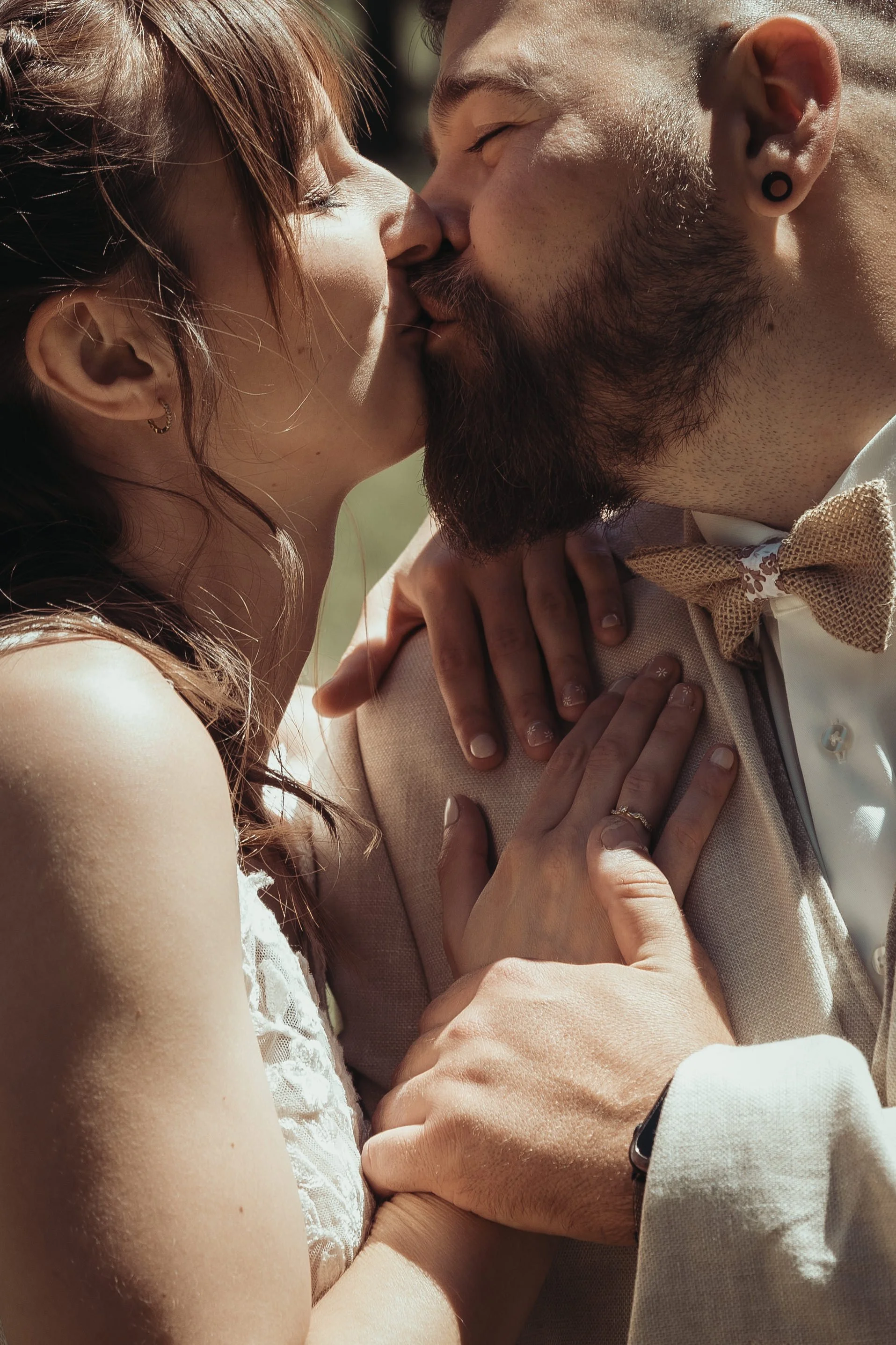 Un couple s'embrasse tendrement, plongé dans un moment intime, lors d'une journée ensoleillée. La femme porte une tenue blanche en dentelle, et l'homme est vêtu d'un costume clair avec un nœud papillon beige. La scène est capturée en gros plan, metta