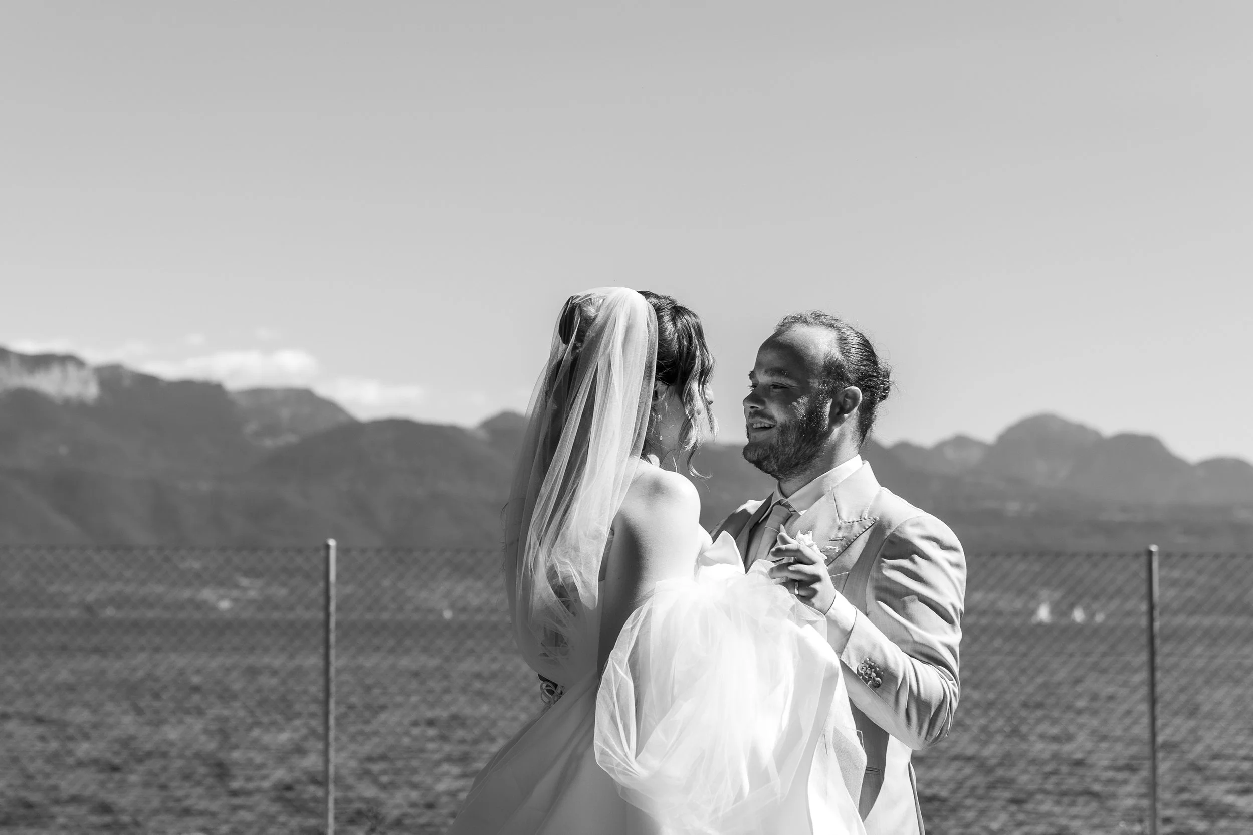 Un couple en habits de mariage souriant, se regardant tendrement, avec une montagne en arrière-plan dans un paysage en plein air.