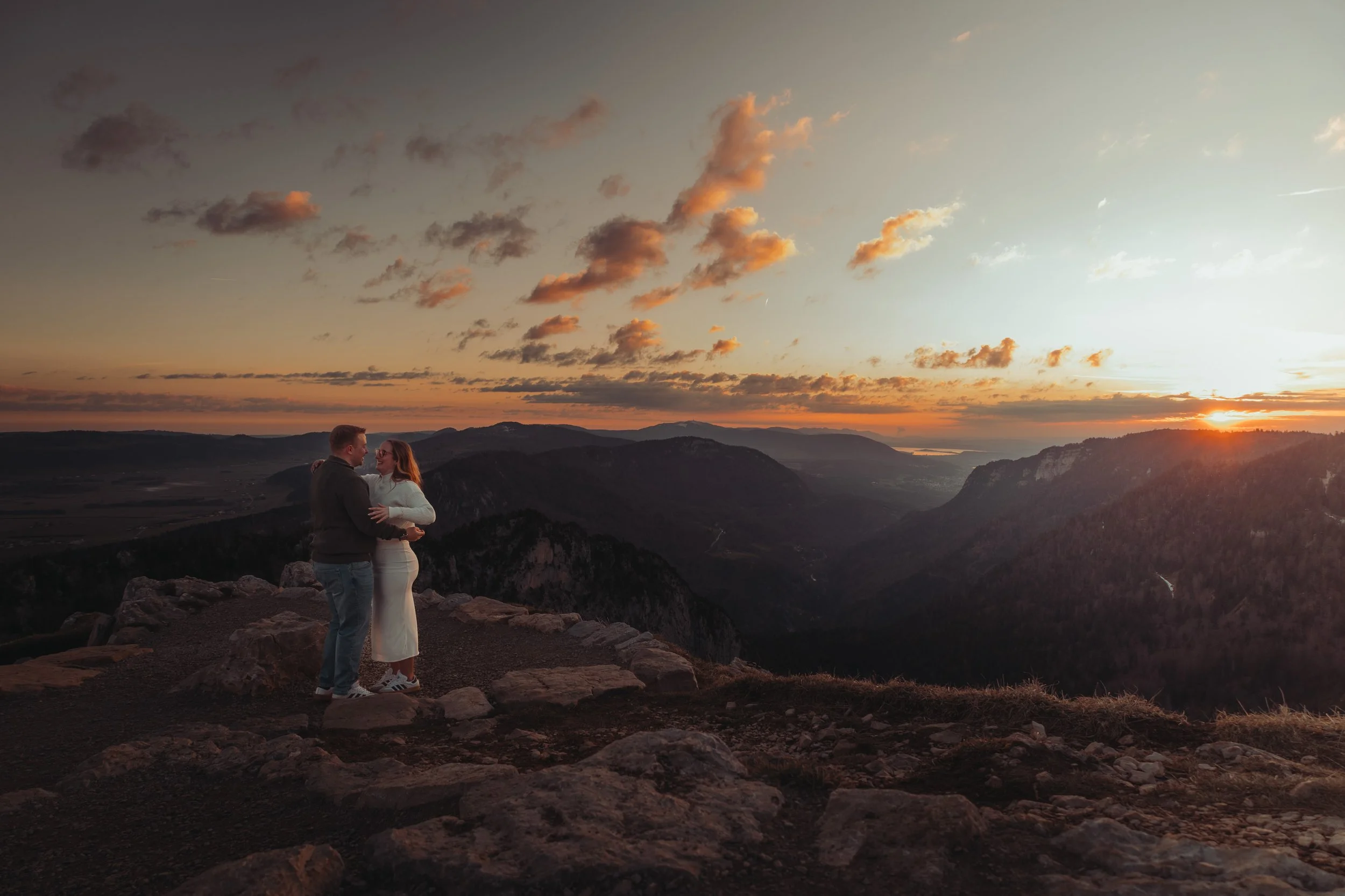 Un couple qui se tient par la taille, regardant dans les yeux, sur une colline au coucher du soleil avec un paysage de montagnes et de lac au loin.