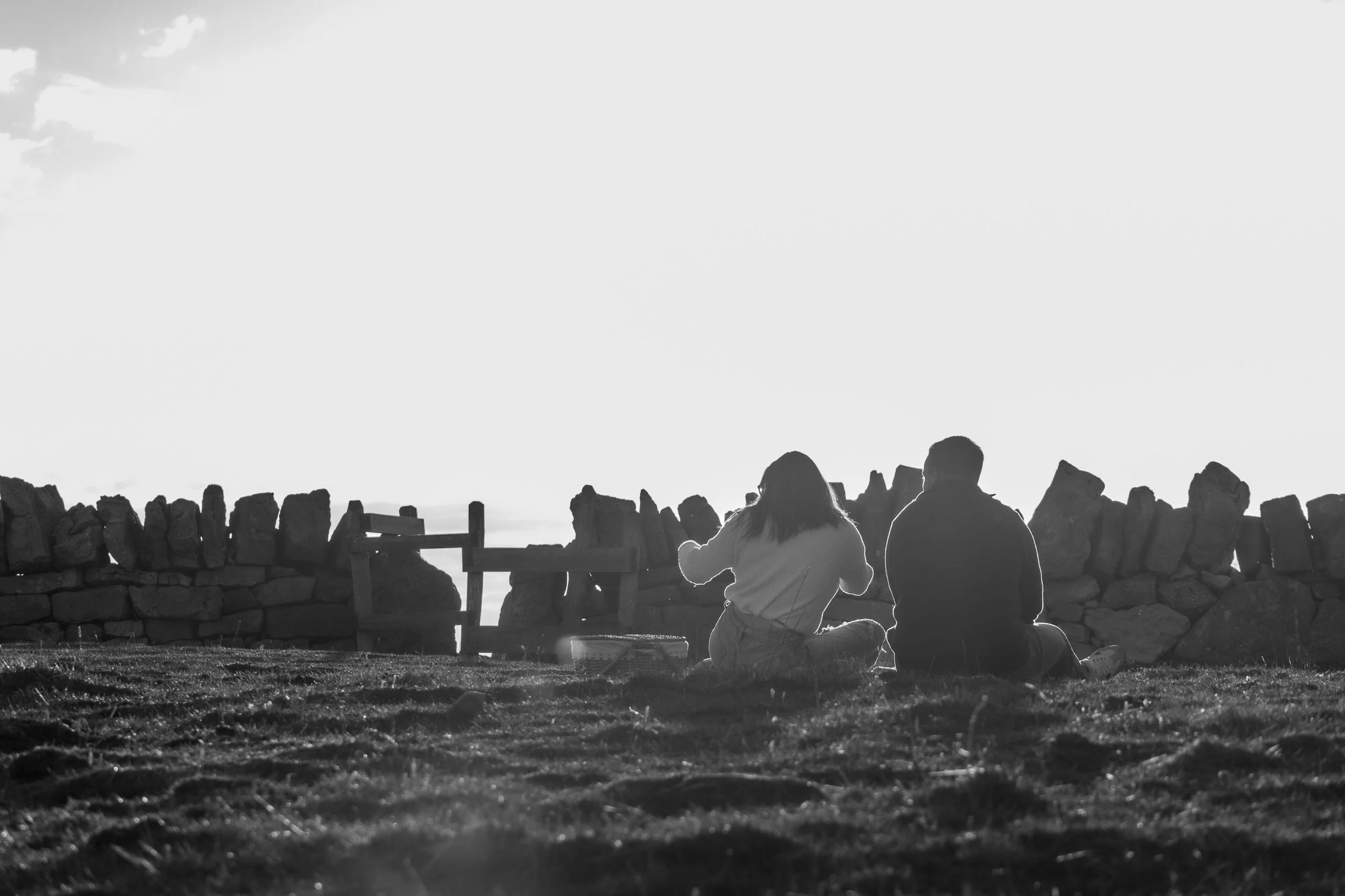 Deux personnes assises sur l'herbe près d'une clôture en pierre, regardant le coucher de soleil.