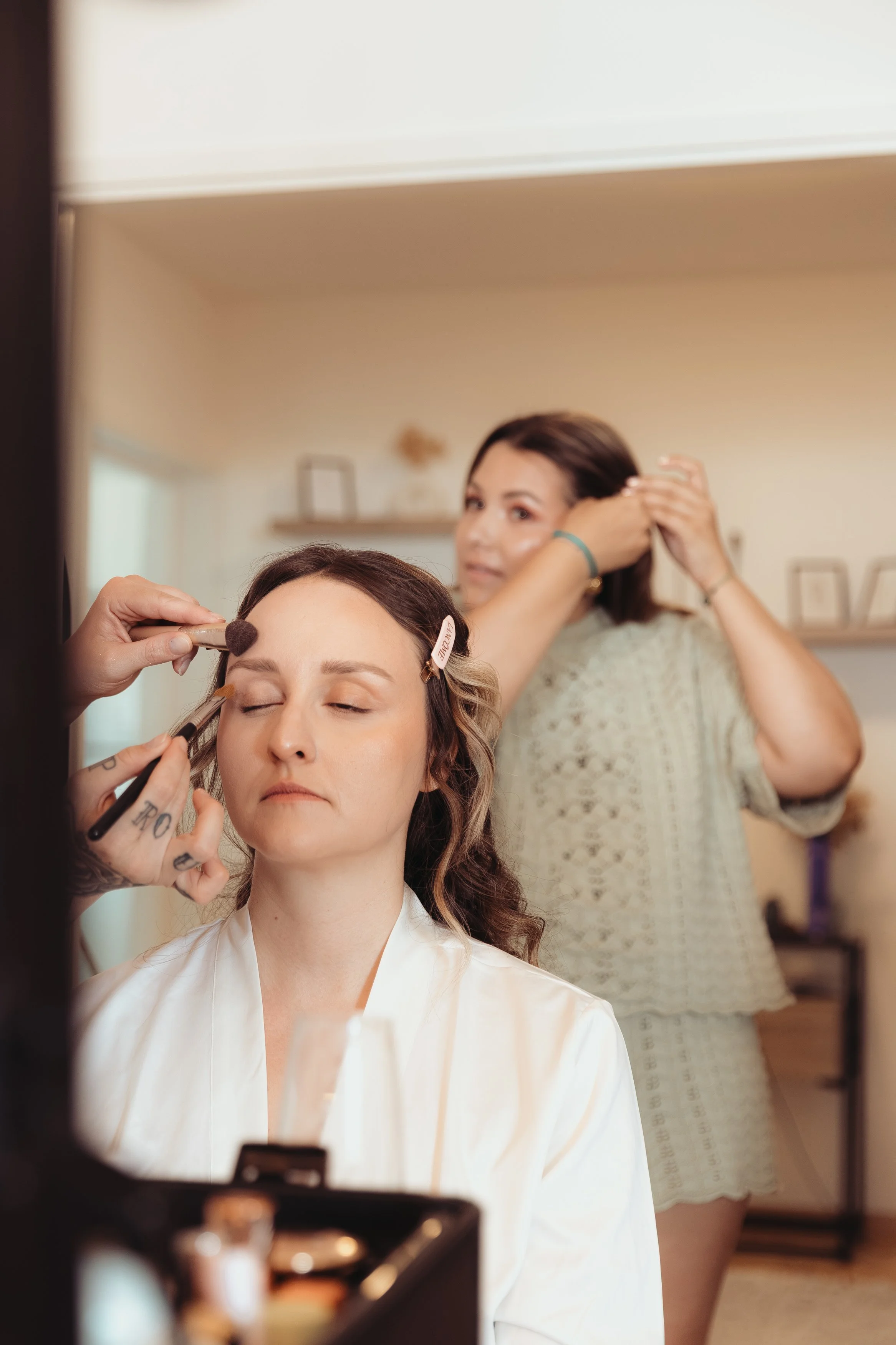 Une femme se fait maquiller par une autre femme dans un salon, en arrière-plan une autre femme se coiffe devant un miroir.