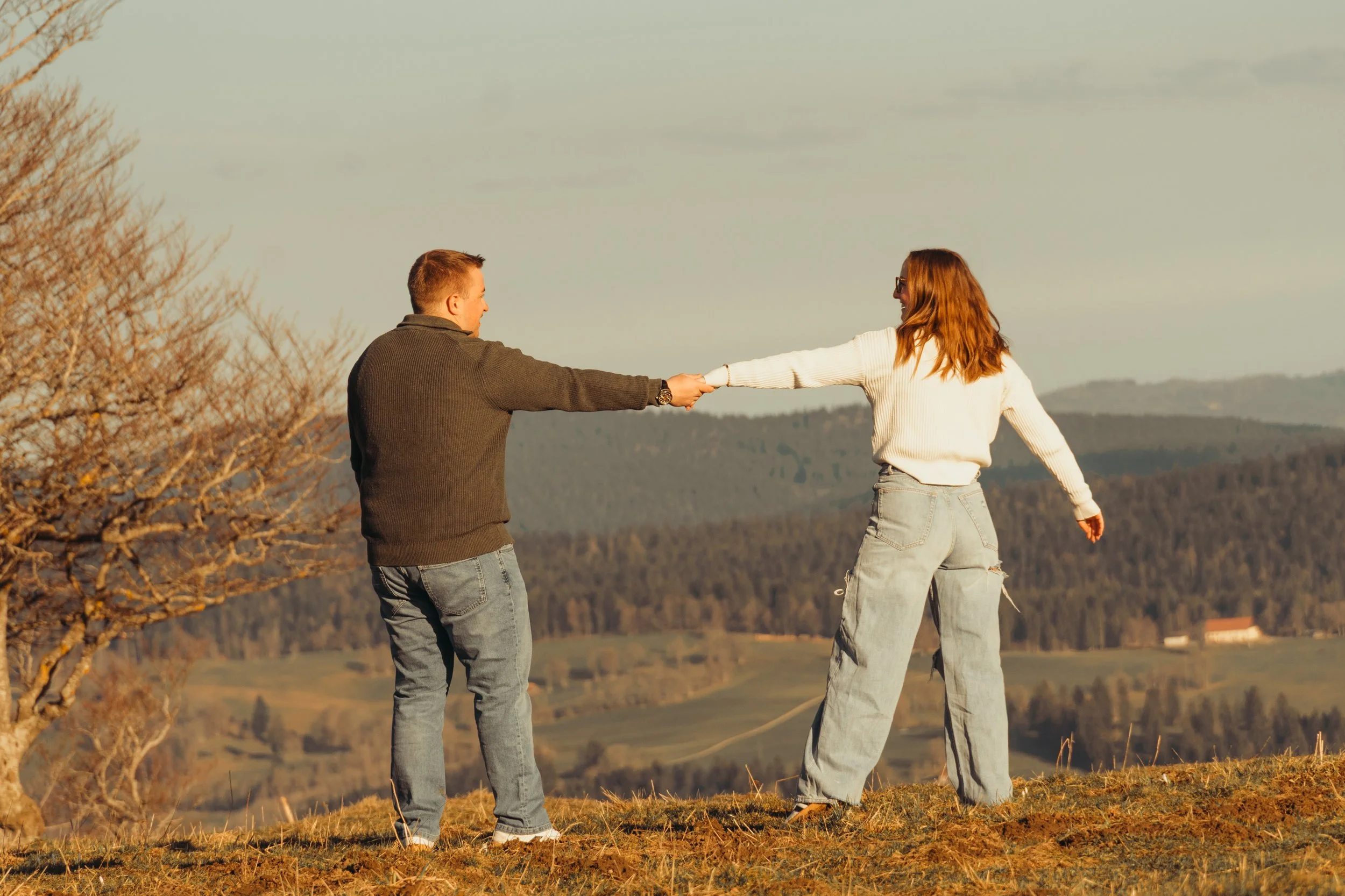 Un homme et une femme se tiennent dans un champ, se tenant la main, avec un paysage rural en arrière-plan, sous un ciel clair.