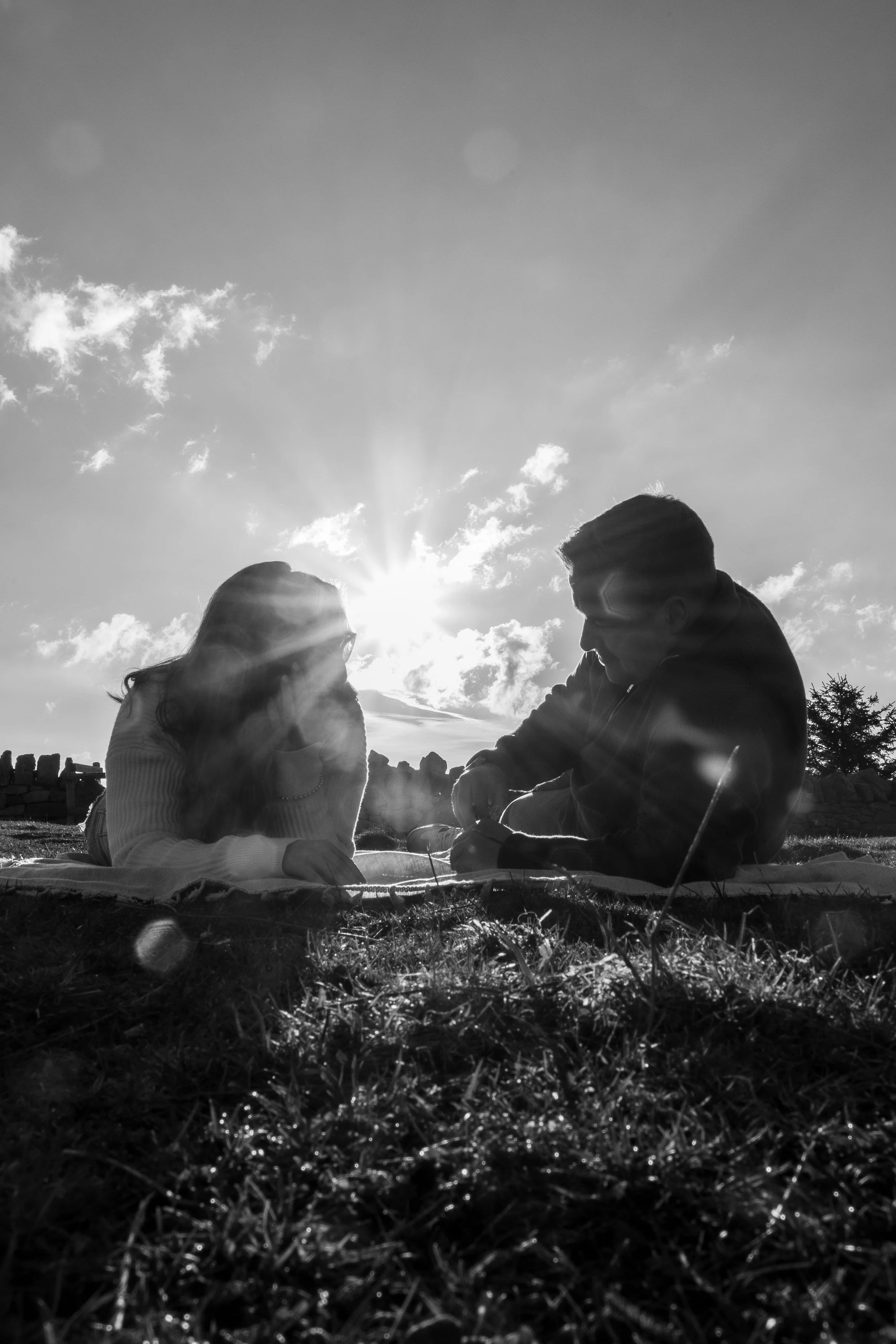 Deux personnes assises sur l'herbe, en silhouette contre le soleil au coucher ou lever du soleil, partageant un moment ensemble.