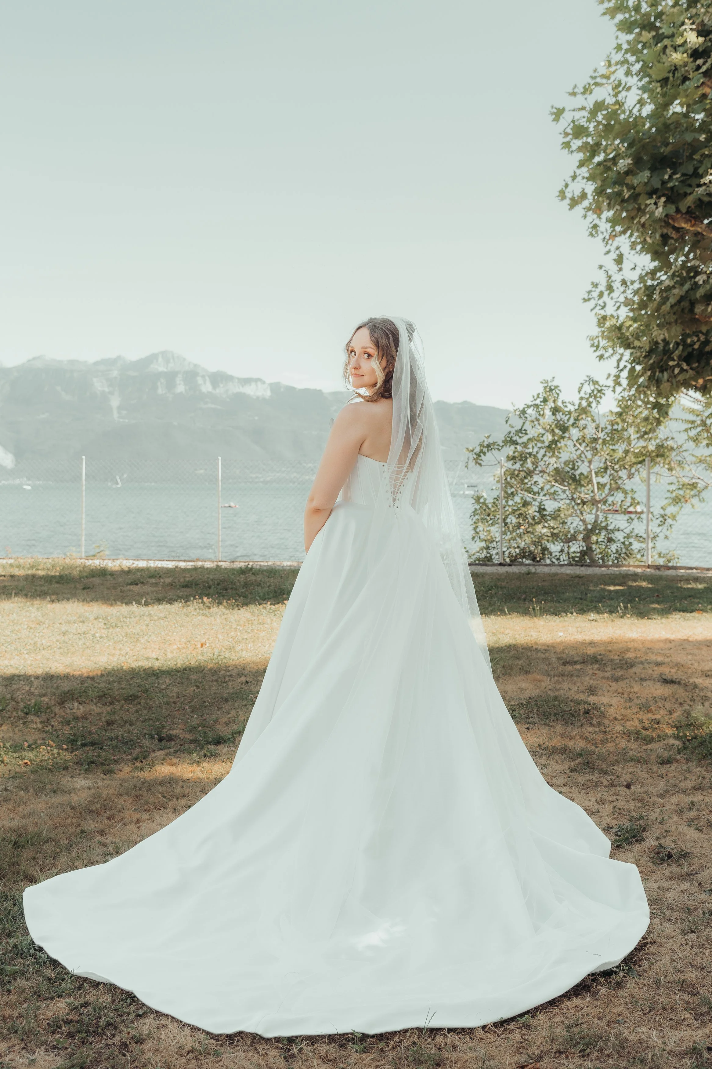 Une femme en robe de mariage blanche avec un voile, debout dans un parc au bord d'un lac, avec des montagnes en arrière-plan et un arbre à droite.