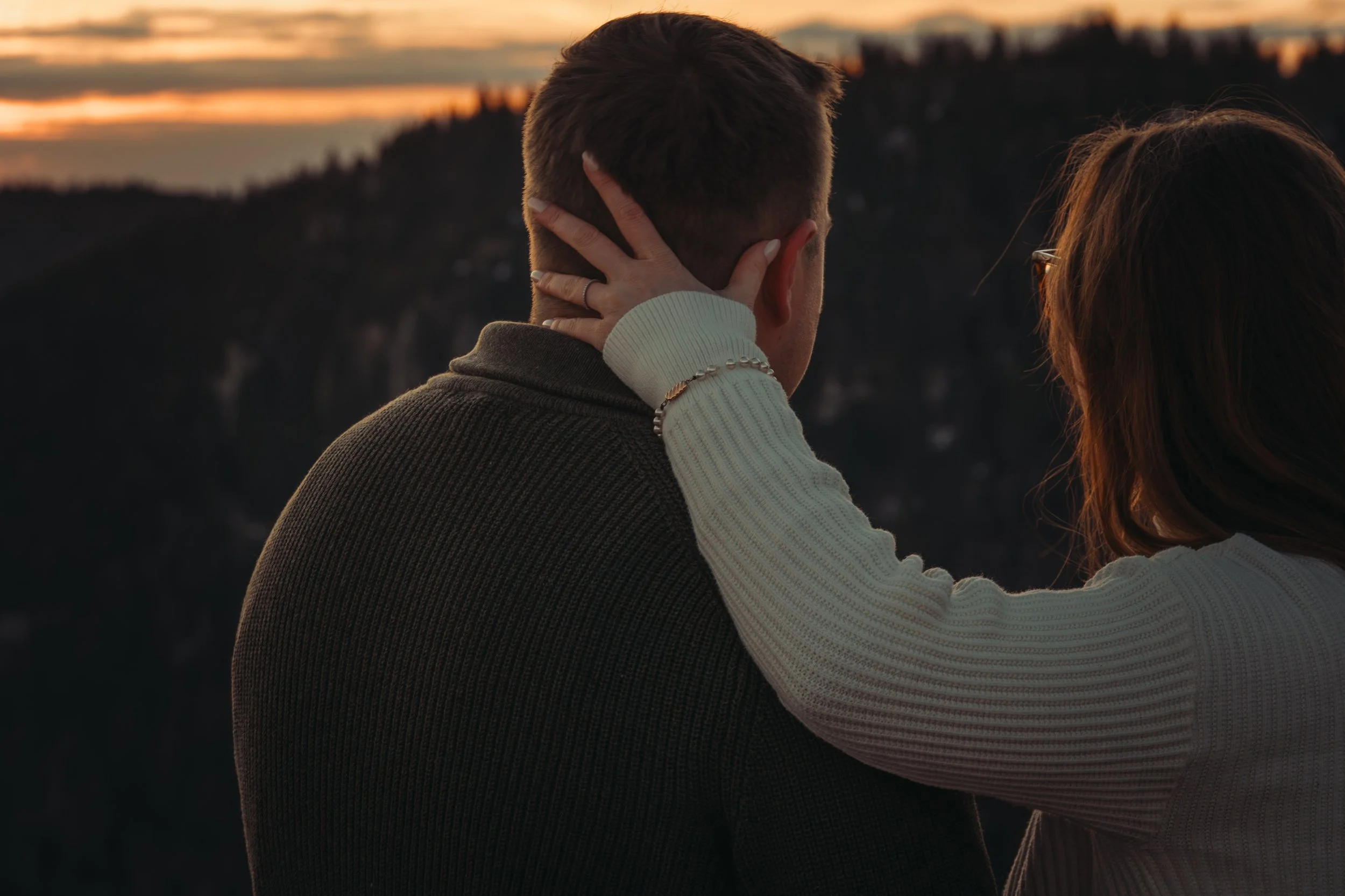 Un couple regarde un coucher de soleil, la femme touche les cheveux de l'homme, dans un environnement naturel.