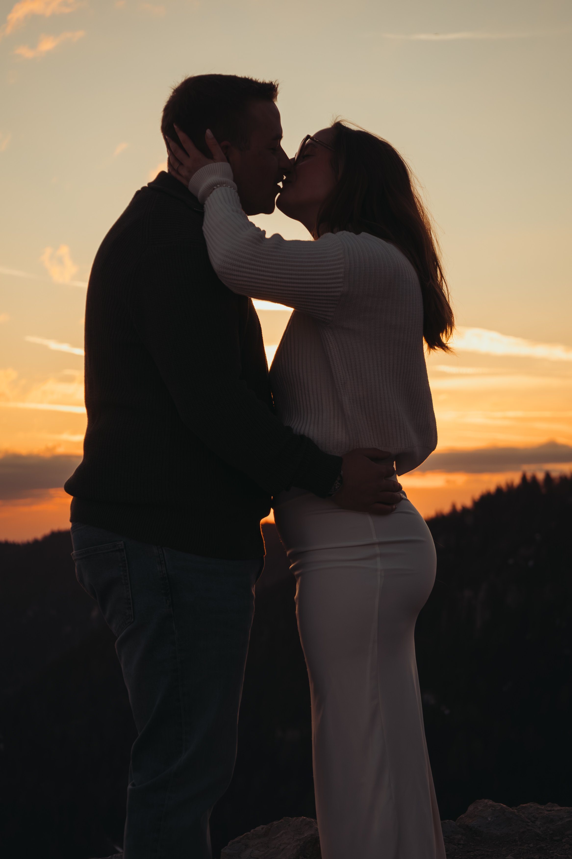 Un couple s'embrasse lors d'un coucher de soleil, silhouette contre un ciel orangé, en plein air, avec des collines en arrière-plan.