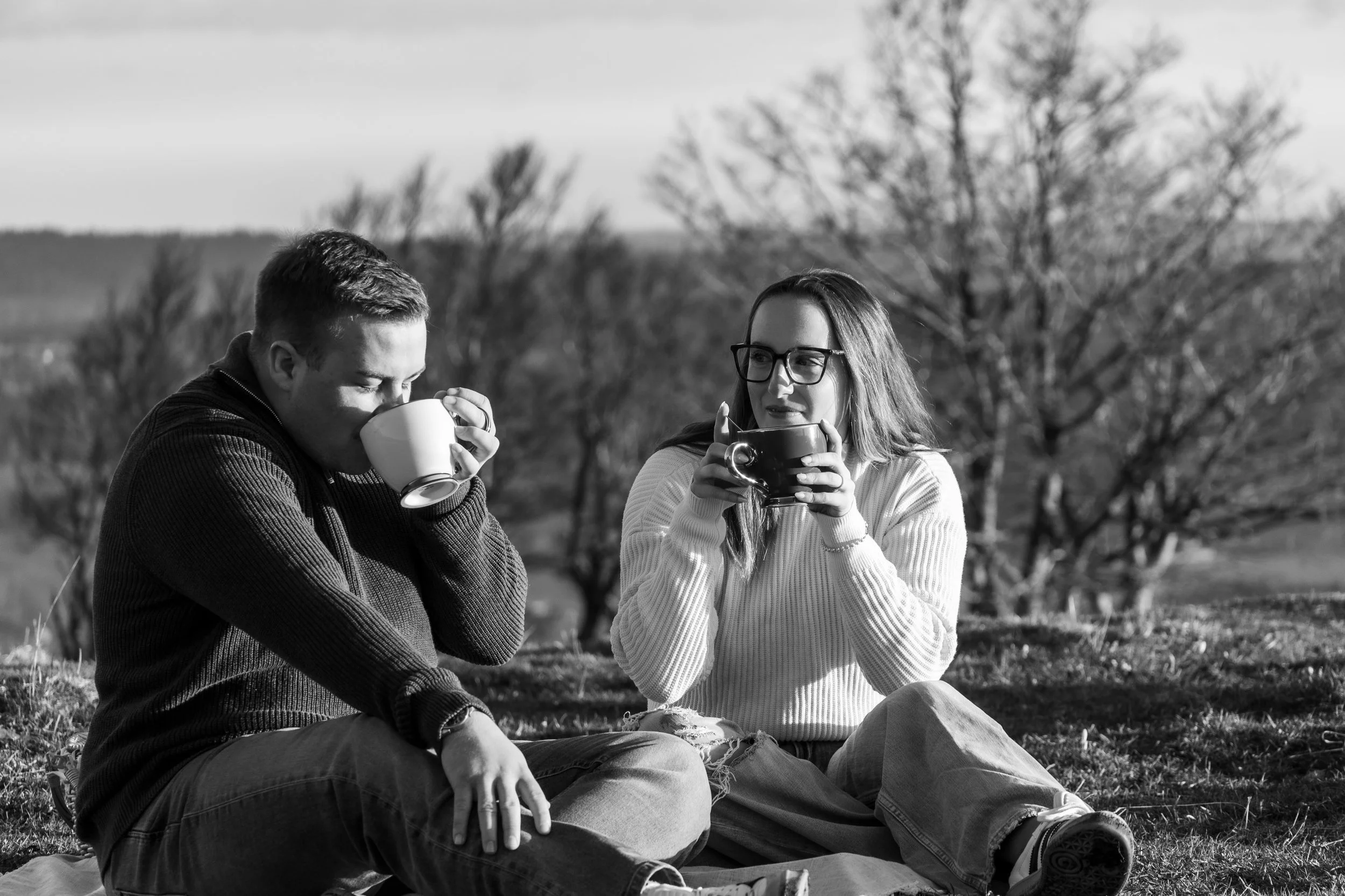 Deux jeunes adultes assis par terre dans un champ, boivent de grandes tasses, en plein air avec des arbres en arrière-plan.
