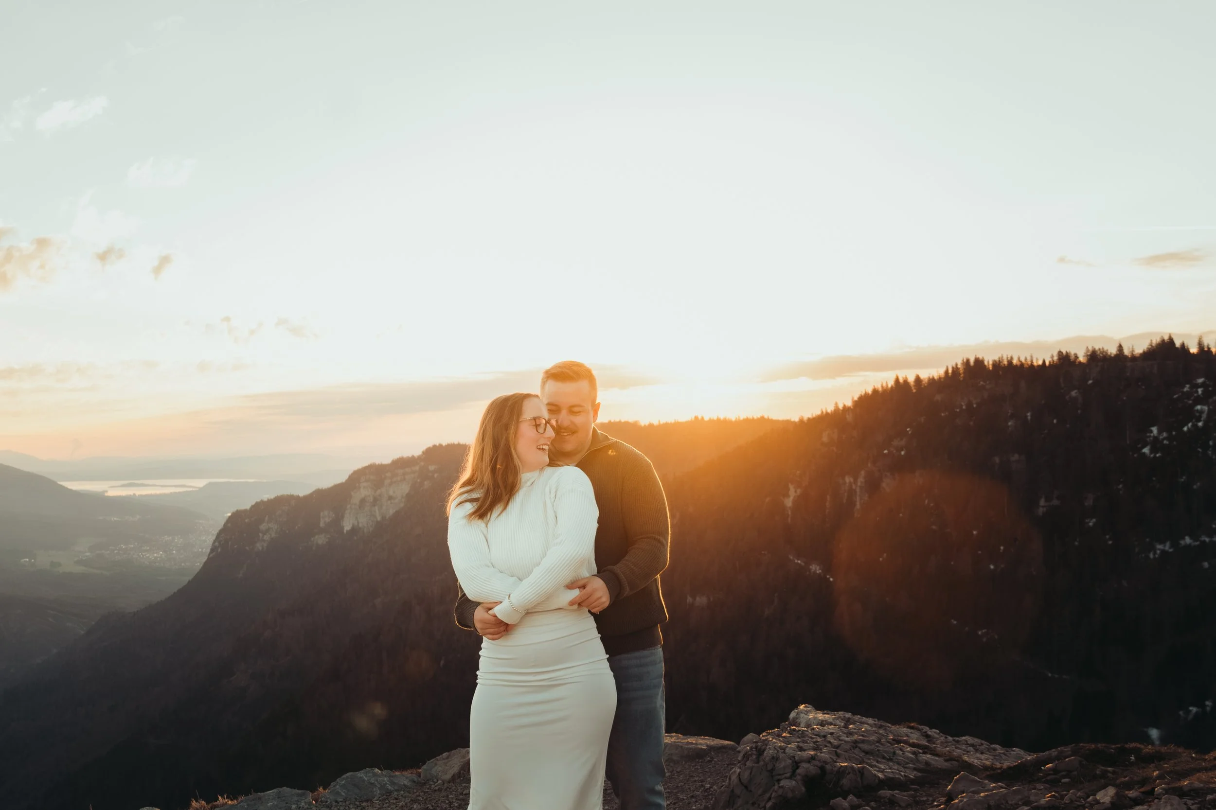 Un couple souriant, main dans la main, lors d'un coucher de soleil dans une région montagneuse avec un lac au fond.
