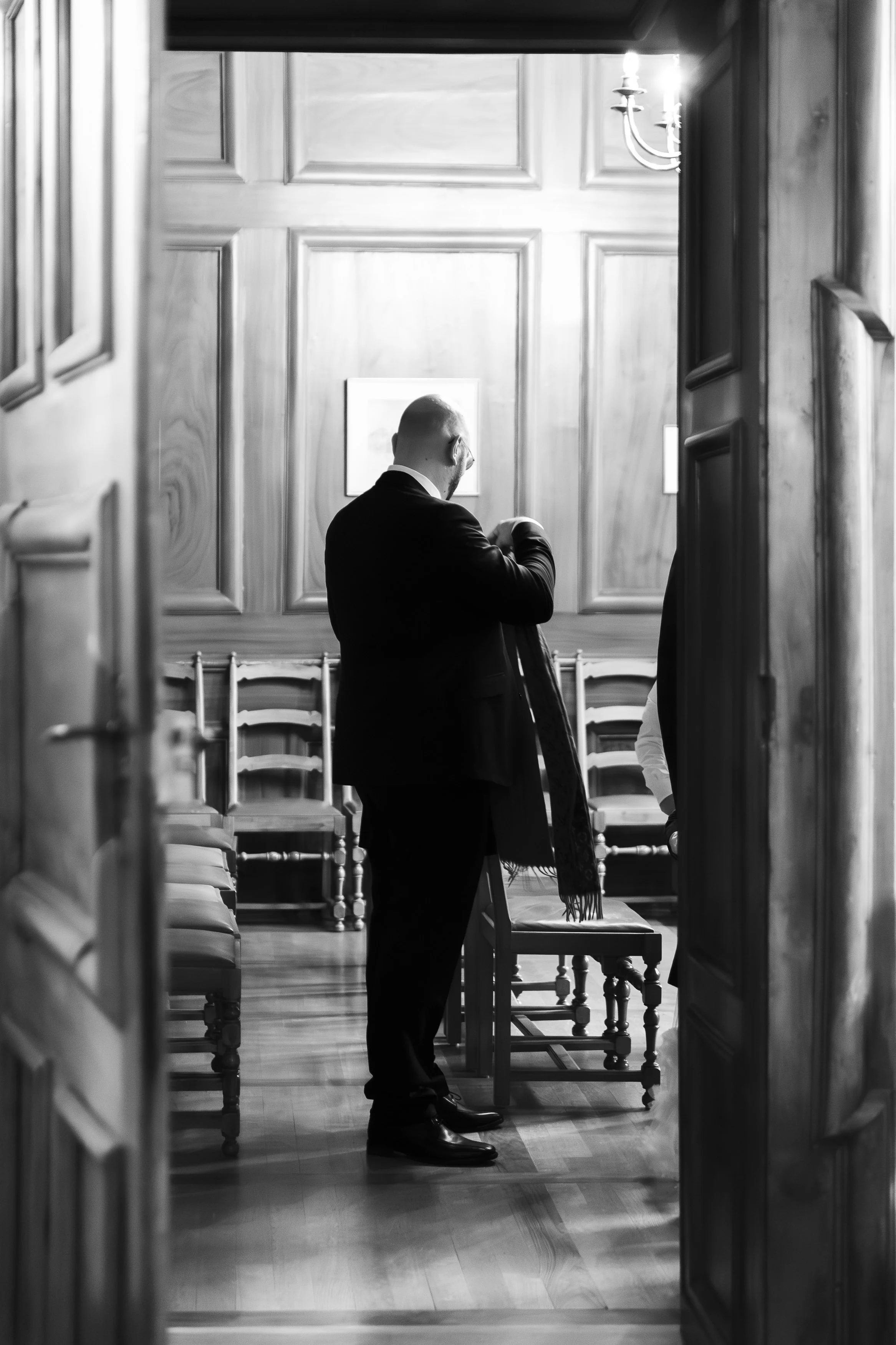 Un homme en costume noir se tient dans une pièce en bois, ajustant une veste ou un manteau, vue partielle depuis une porte entrouverte, dans un cadre élégant en noir et blanc lors d'un reportage mariage avec Marie Monod photographe d'émotions