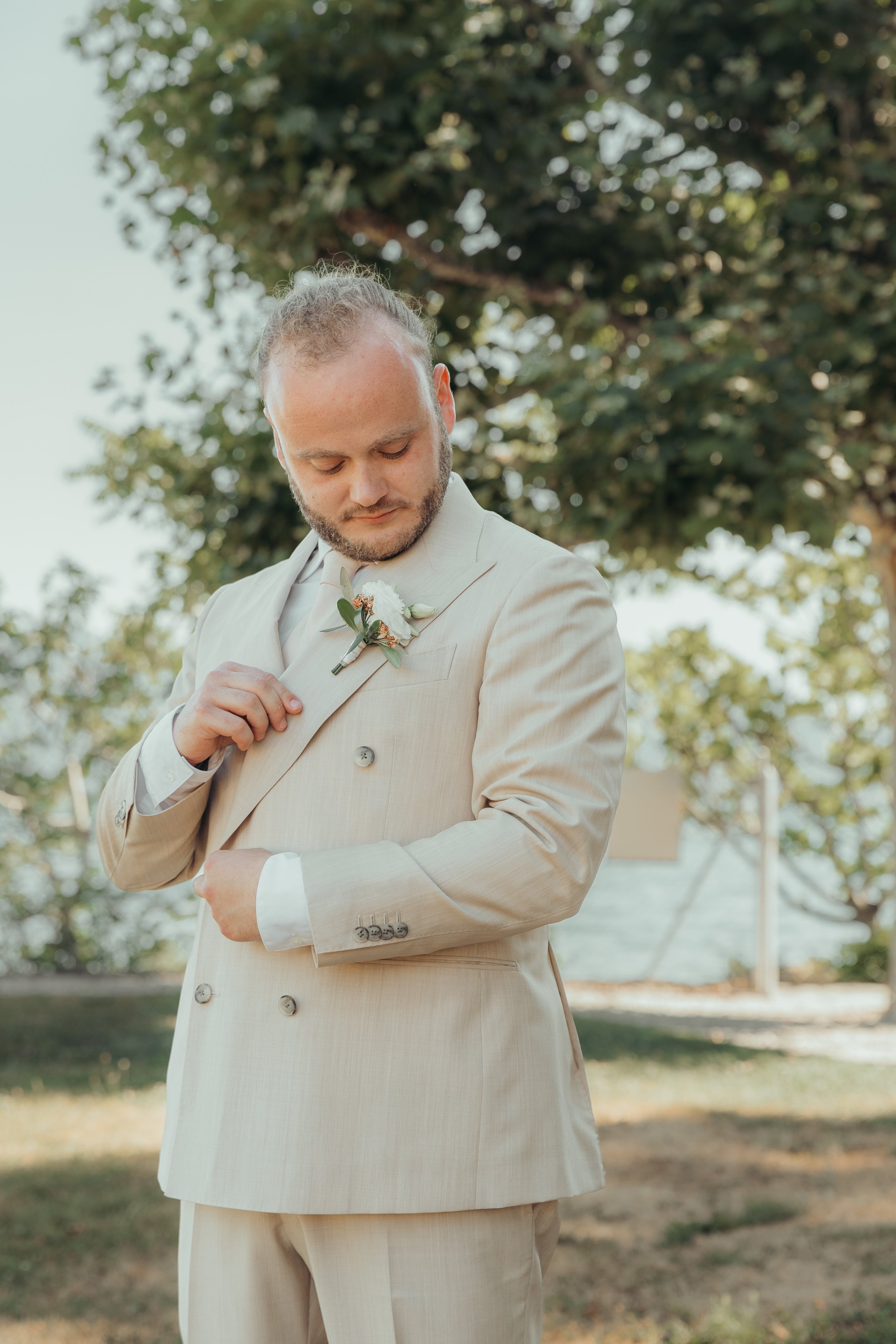 Un homme habillé d'un costume beige en extérieur, ajustant la boutonnière de sa veste, avec arbres en arrière-plan.