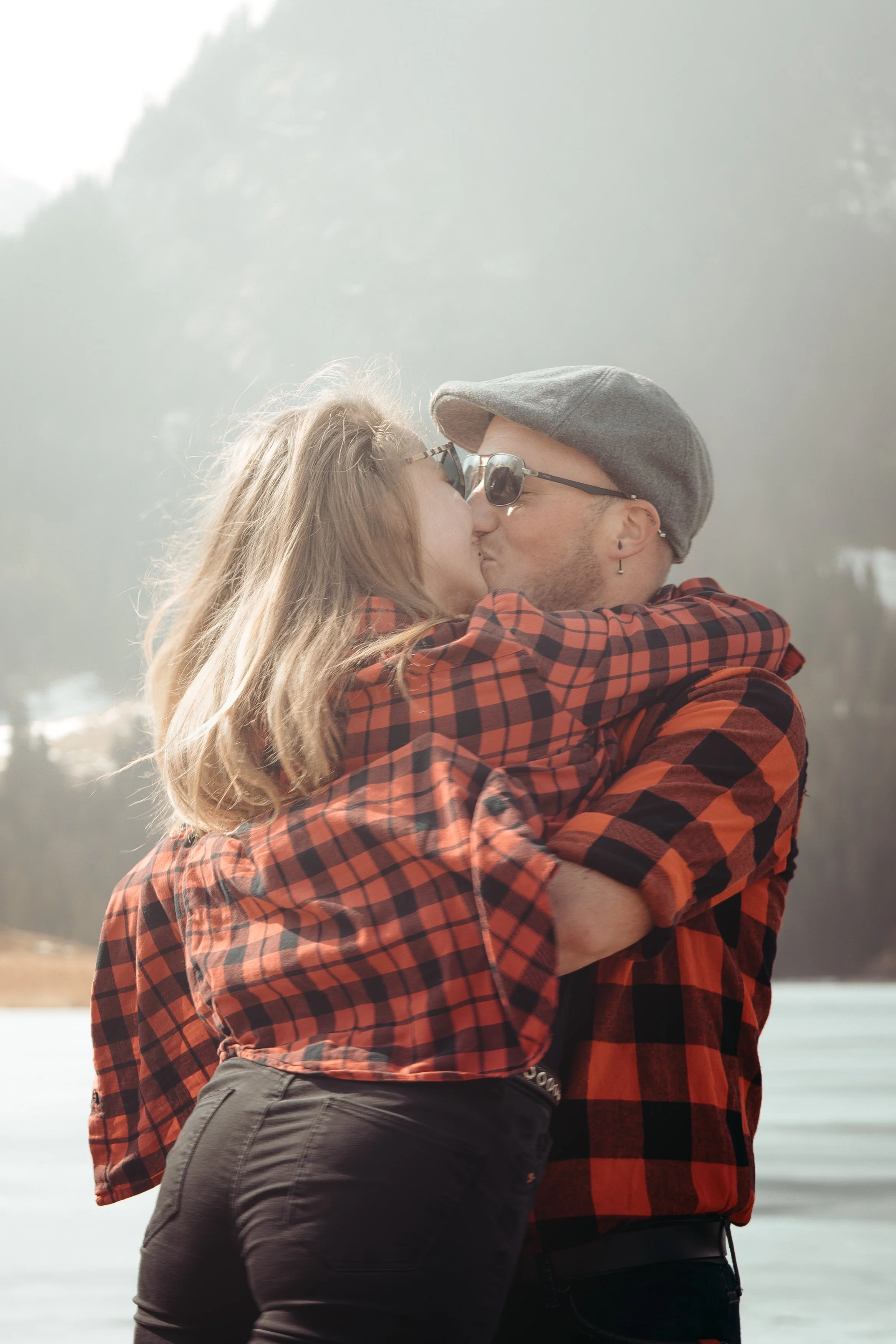 Un couple s'embrasse en plein air dans un environnement naturel, portant des chemises à carreaux rouges et noirs, avec un paysage de montagne brumeuse en arrière-plan.