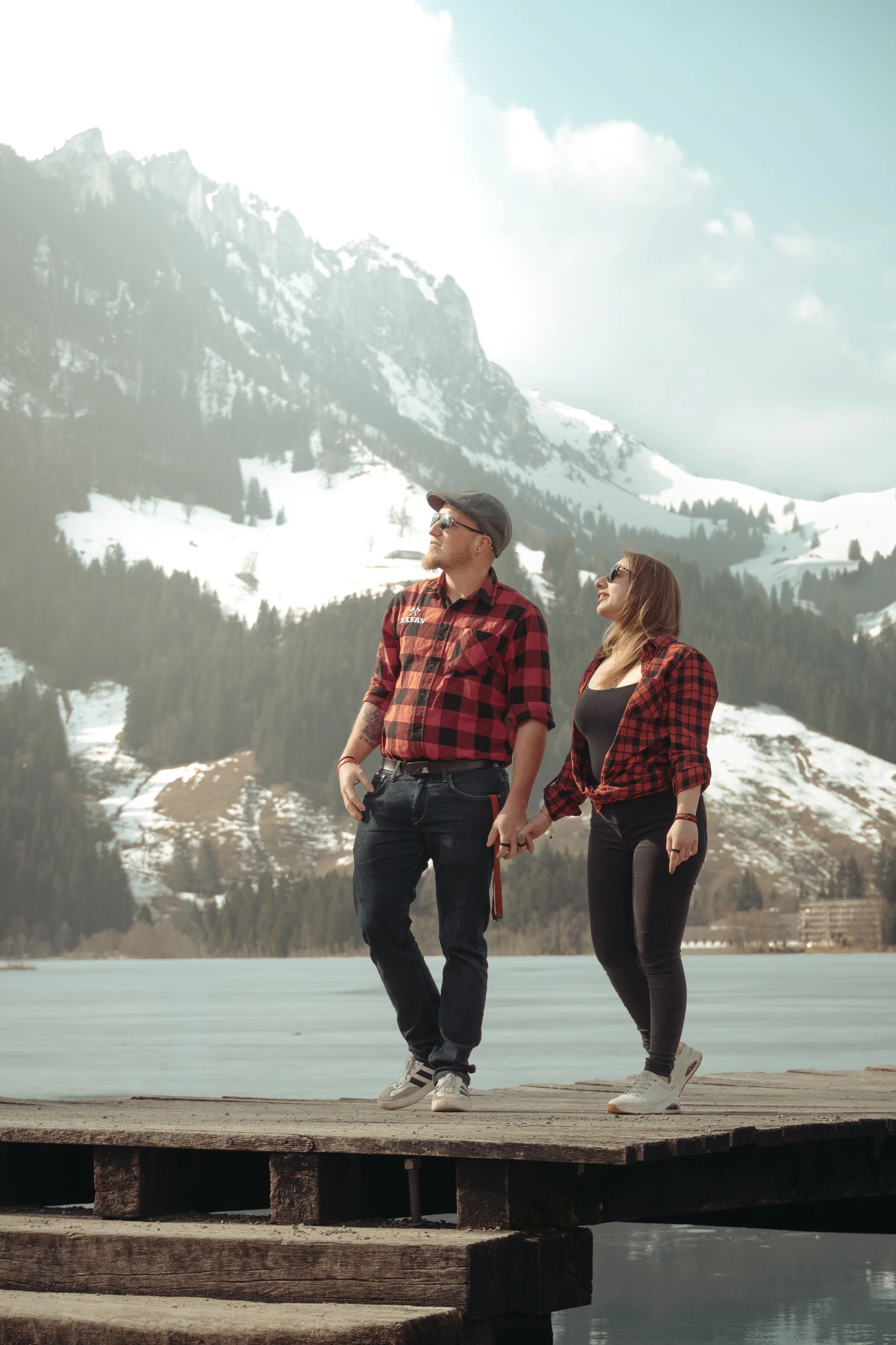 Un couple tenant la main, habillé en chemises à carreaux rouges, se promène sur une jetée en bois au bord d'un lac, avec des montagnes enneigées en arrière-plan.