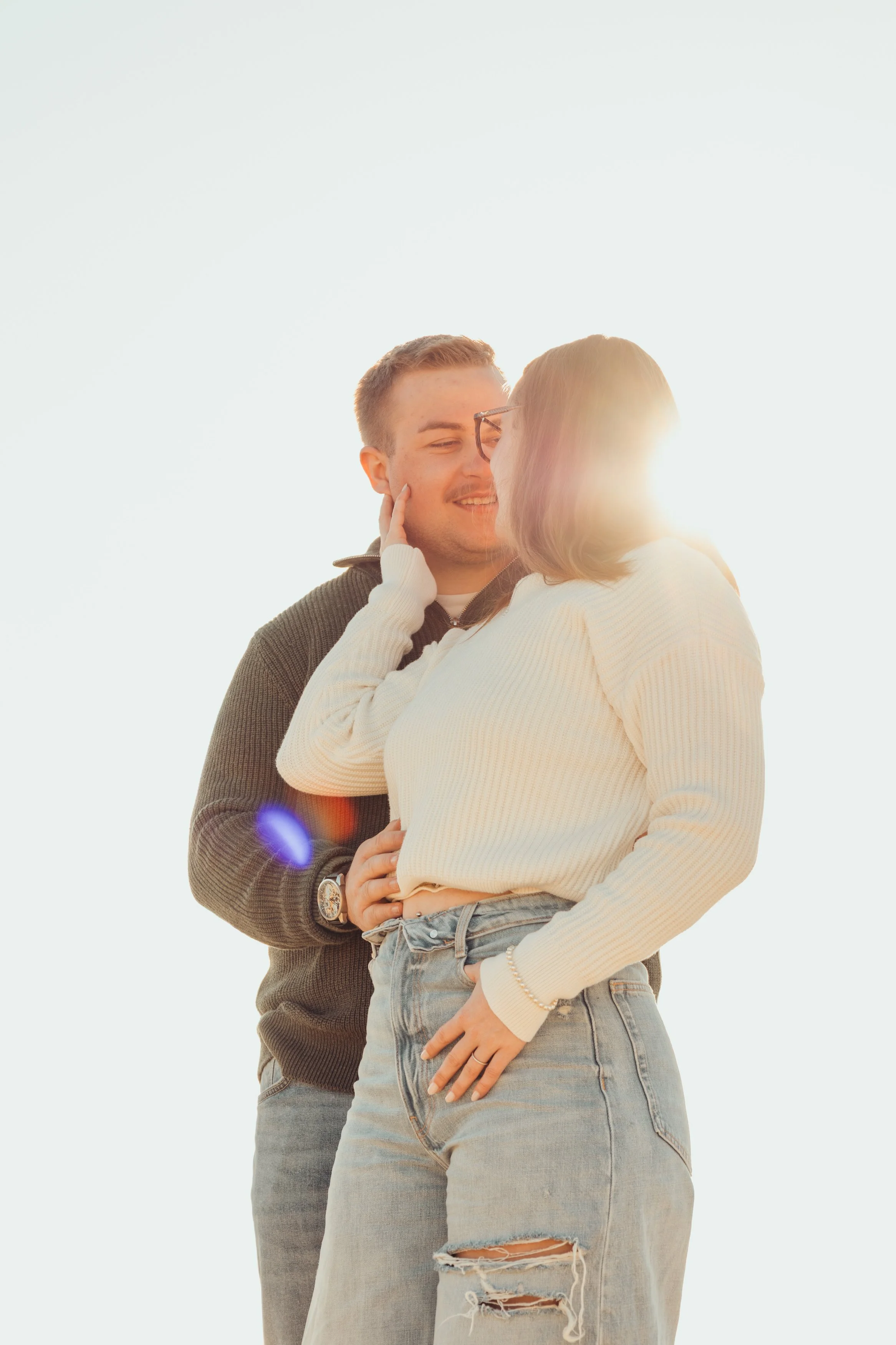 Un couple s'embrasse dans un extérieur ensoleillé, avec le soleil derrière eux créant un éclat de lumière. La femme porte un pull blanc et un jean déchiré, tandis que l'homme porte un pull gris foncé ou marron clair et un jean. Ils semblent très proc