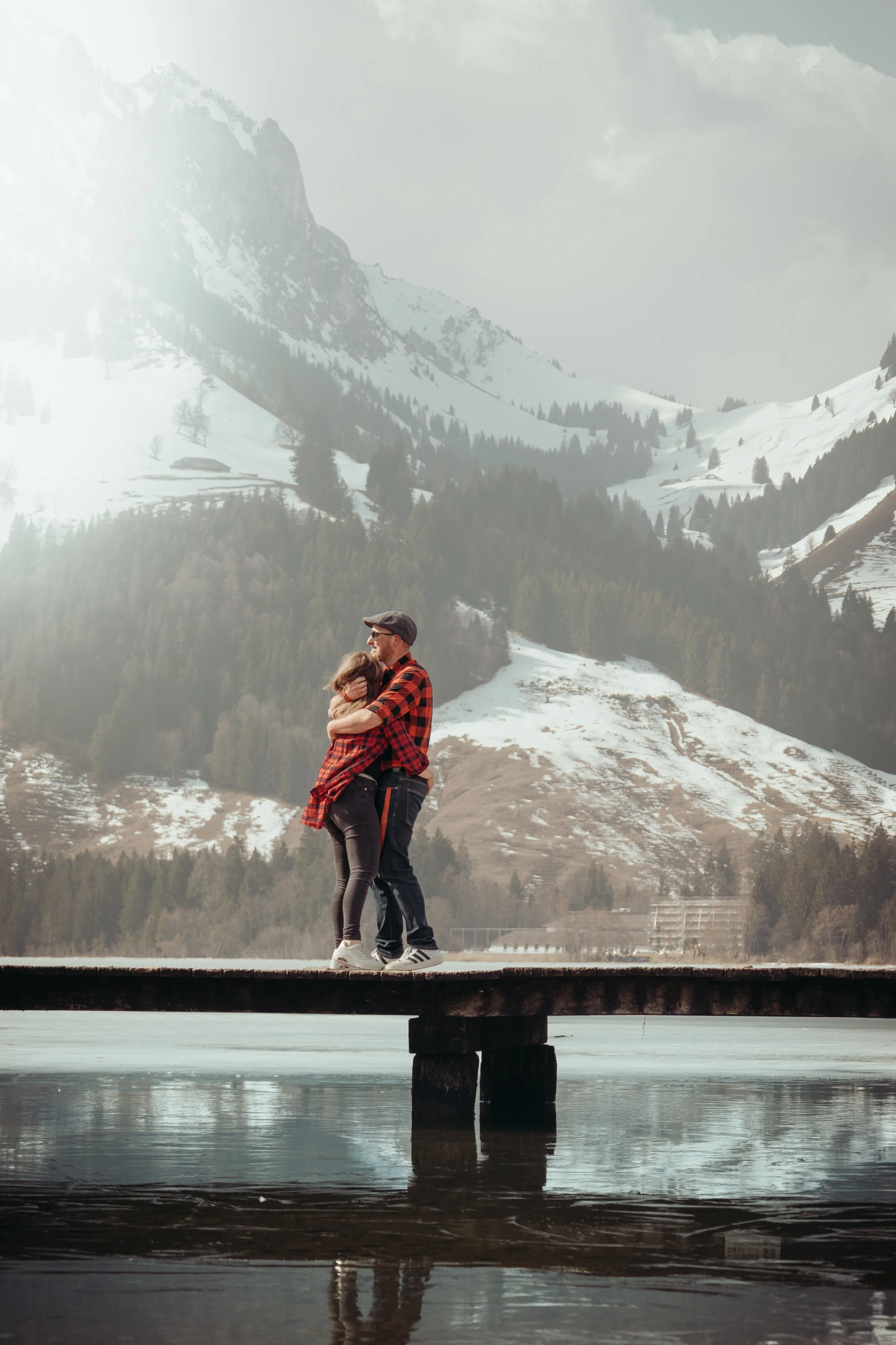 Un couple qui s'embrasse sur un pont en bois au bord d'un lac, avec des montagnes enneigées en arrière-plan.