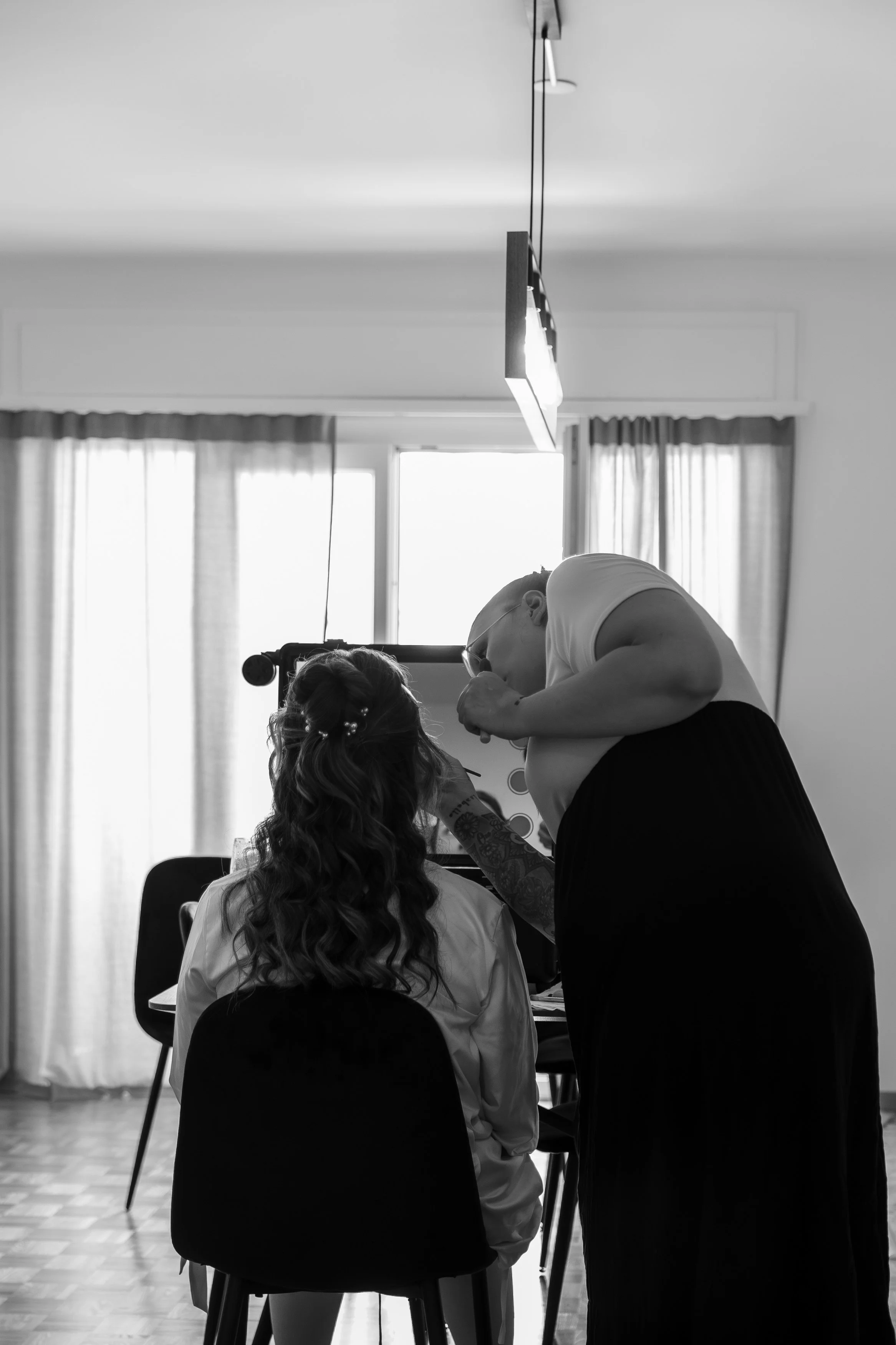 Une femme assise voit un professionnel effectuer un maquillage ou une application de maquillage sur son visage dans une pièce bien éclairée, avec des rideaux devant une fenêtre.